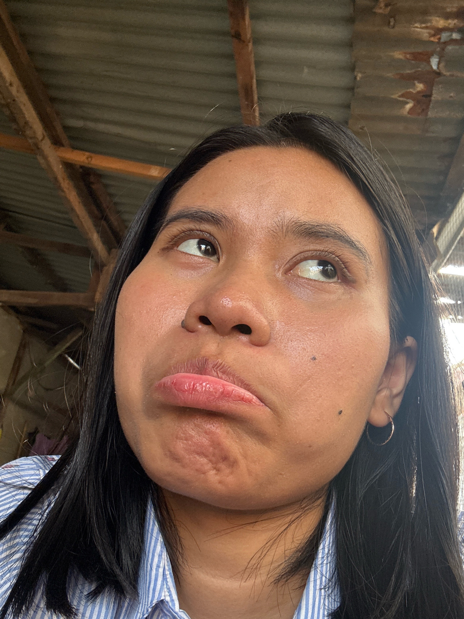A medium shot shows a young woman with dark hair and brown eyes, making a pouty face as she looks up and to her right. Her skin is tan with some blemishes. She is wearing a blue and white striped shirt and a small gold hoop earring in her right ear. The background appears to be an outdoor setting with a corrugated metal roof and wooden beams, suggesting a rural or construction area. The image was taken in East Jakarta, Indonesia.
