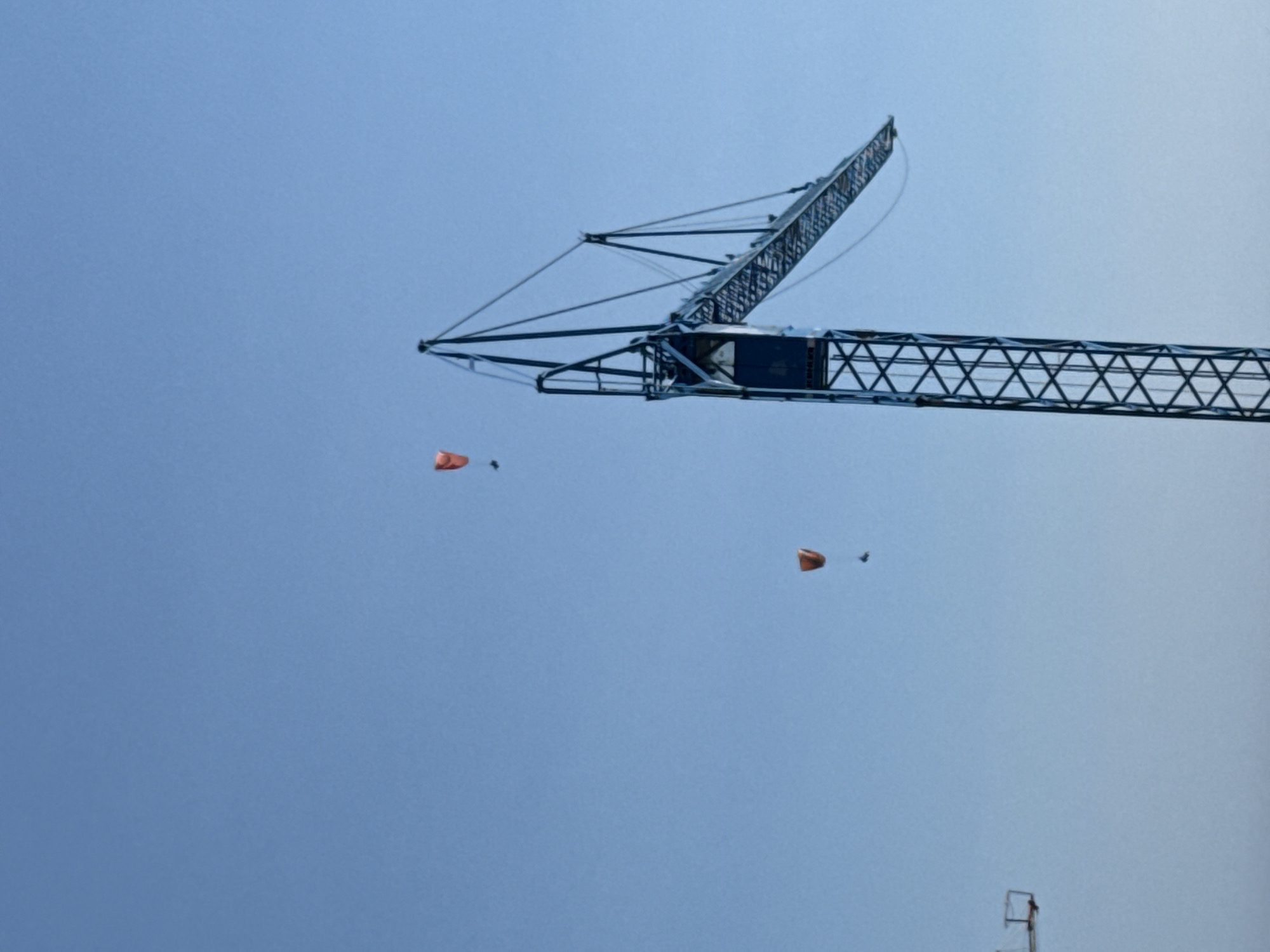 In Boreti, Montenegro, two individuals are paragliding against a clear blue sky. One paraglider, with an orange wing, is positioned to the left of a large blue construction crane. The second paraglider, also with an orange wing, is higher and to the right of the first, and also to the left of the crane. The construction crane dominates the right side of the image, its boom extending towards the right. A portion of another, less distinct construction structure is visible on the far left of the image.