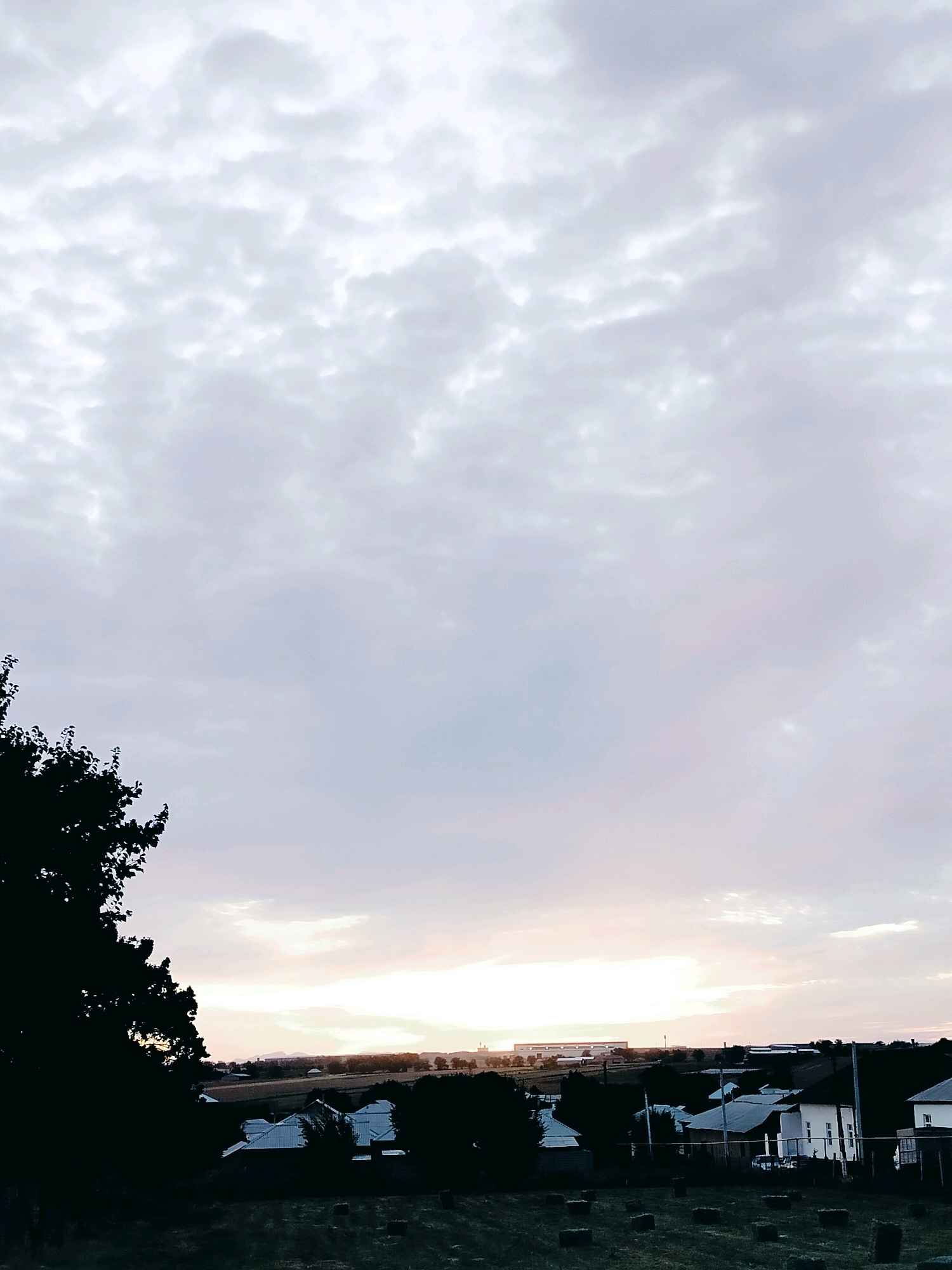 The image shows a rural landscape at dusk, likely in or near the city of Көлкент, Kazakhstan. A large area of overcast sky dominates the upper portion of the frame, with soft, diffused light indicating the setting sun behind the clouds near the horizon. In the foreground, silhouetted trees on the left frame the scene. Below the sky, a settlement of houses with white roofs is visible amidst darker foliage. The houses appear to be single-story structures with a few windows discernible. In the field in front of the houses, several rectangular hay bales are scattered. In the distance, beyond the settlement, a long, low-rise building or complex is visible, possibly an industrial or agricultural facility, and a low mountain range can be seen on the far horizon. The overall atmosphere is serene and quiet, typical of a rural evening. There are no people or animals visible in the image.