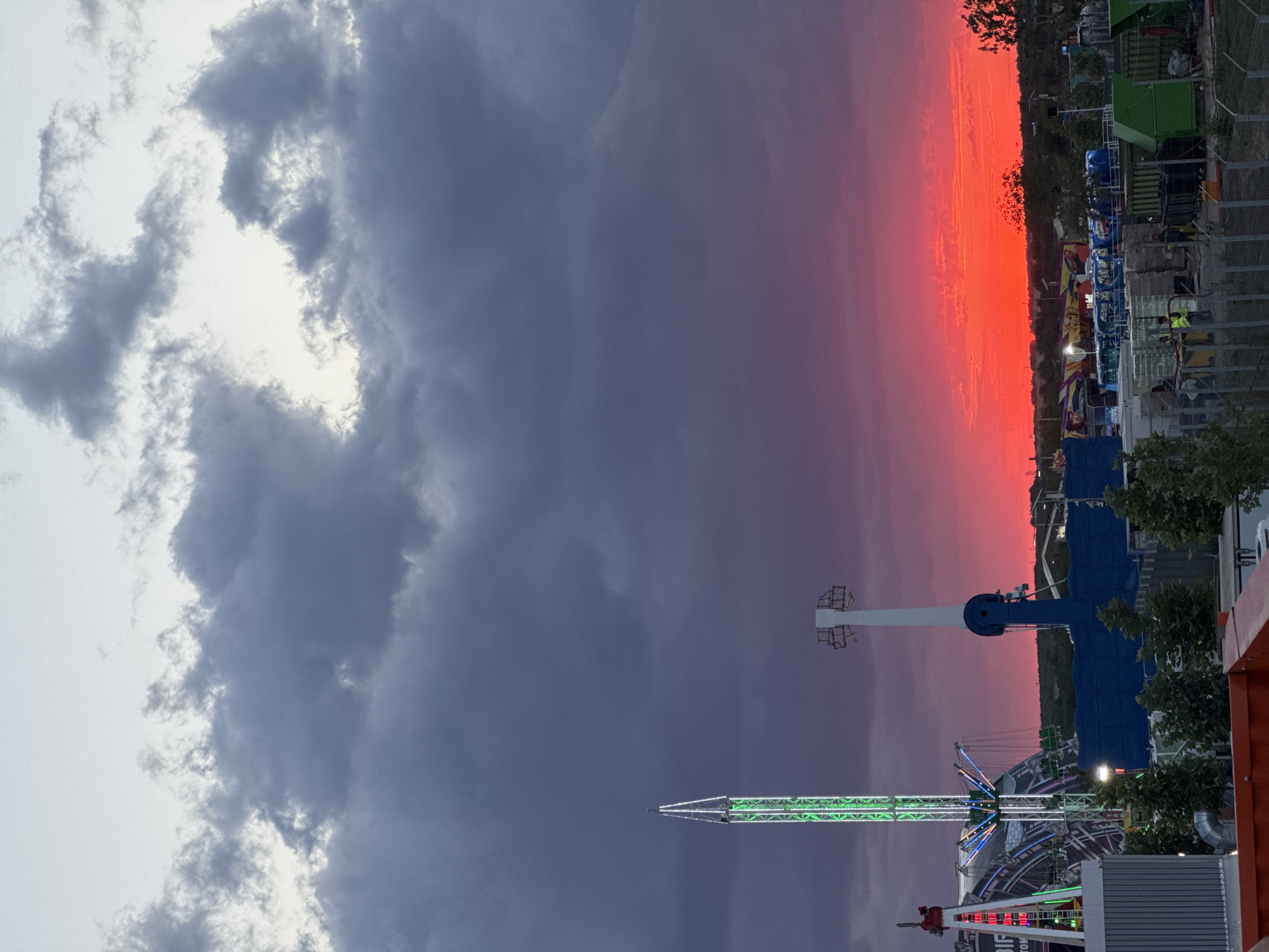 The image depicts an amusement park with a dramatic sunset over the city of Costinești, Romania. The sky is a vibrant spectacle of deep purple clouds transitioning to a fiery orange and red horizon. Prominent amusement park rides, including a tall green tower ride illuminated with lights and a white pendulum ride, dominate the mid-ground. In the foreground, various structures and equipment, possibly related to the park's operations or ongoing construction, are visible. The overall scene captures the lively atmosphere of the amusement park set against a breathtaking natural phenomenon.