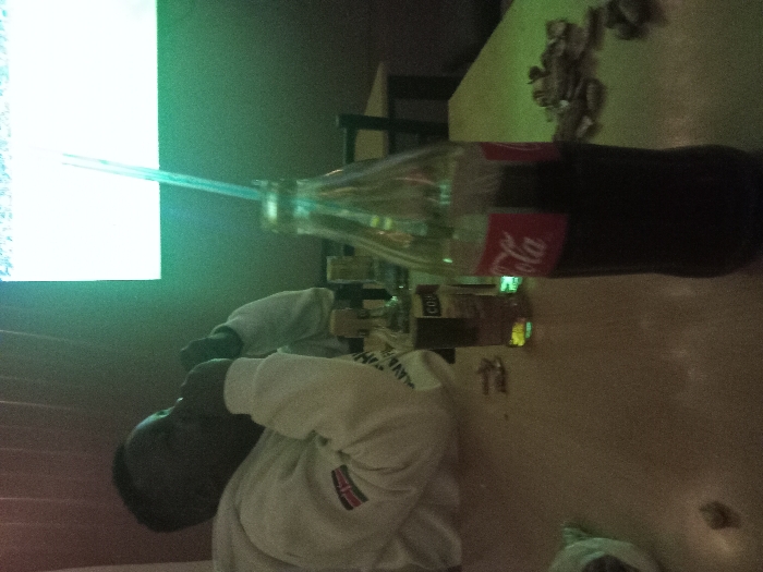 A person in a white sweater with a Kenyan flag patch drinks a Coca-Cola bottle using a straw. The bottle is tilted on a table, and several small objects are scattered nearby. A bright screen glows in the background.