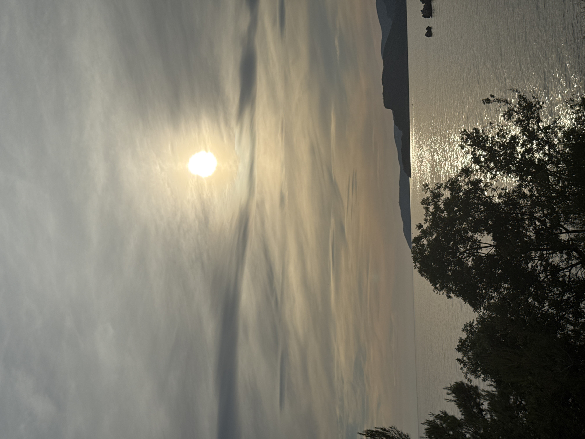 The image depicts a serene seascape with the sun shining brightly through a cloudy sky. In the foreground, silhouetted trees frame the view of the water. The sea is calm, reflecting the sunlight and creating a shimmering effect. In the distance, rolling hills or mountains form the coastline. The location is identified as Sveti Stefan, Montenegro, suggesting a view of the Adriatic Sea from this picturesque coastal area. The scene is devoid of any people or overt human activity, focusing solely on the natural landscape and atmospheric conditions.