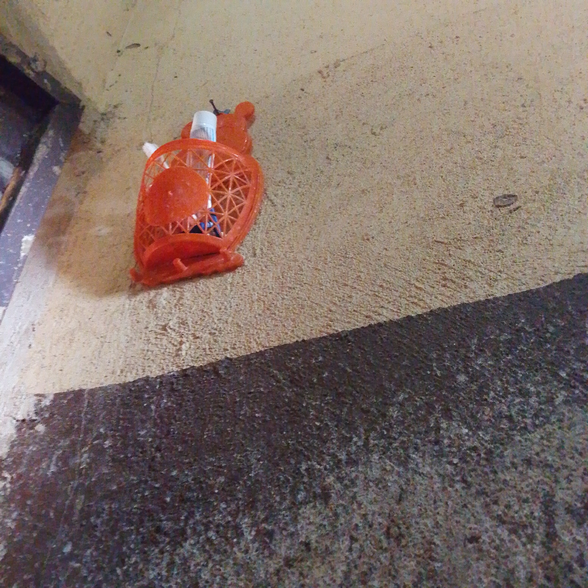A close-up, low-angle shot captures an orange plastic basket, possibly a toy, resting on a textured, light-colored wall. A white plastic object with a blue stripe is visible inside the basket. The basket is positioned in the corner where the light-colored wall meets a darker, rougher surface that slopes diagonally across the lower portion of the frame. This area is located in Jalingo, Nigeria. The lighting appears to be natural, casting a slight shadow from the basket onto the wall. The wall itself shows signs of age and wear.