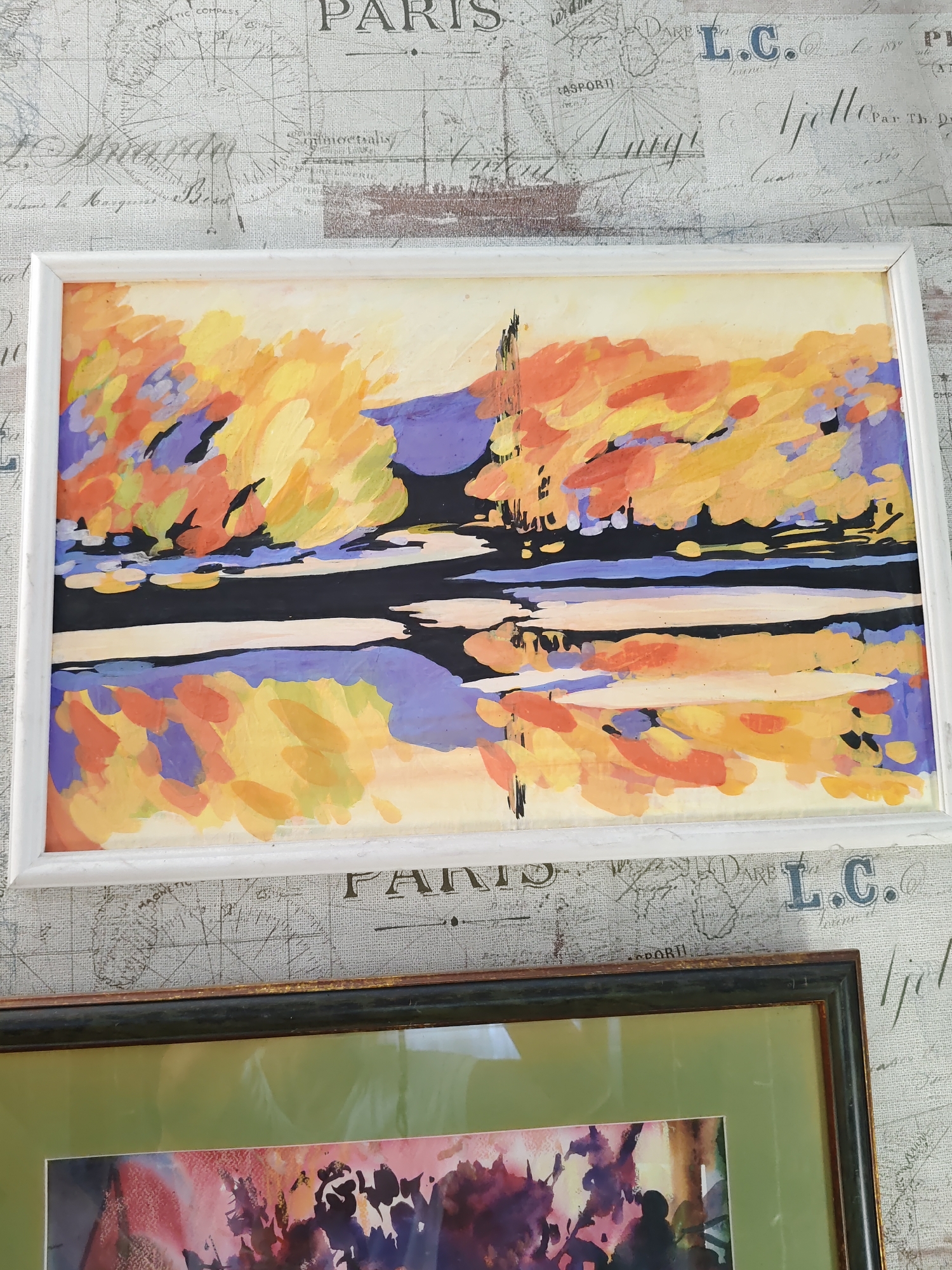The image shows two framed artworks hanging on a wall with a patterned wallpaper. The top artwork is a landscape painting in a white frame, featuring abstract, brushstroke-like representations of trees with vibrant yellow, orange, and purple foliage, reflected in dark water. A thin, dark vertical line, possibly a tree or a pole, stands out against the colorful background. The bottom artwork is partially visible, framed in a dark, textured frame with a light green mat. It appears to be another painting, with abstract shapes in shades of purple, pink, and yellow. The wallpaper behind the artworks has a vintage map and script design. There is no indication of the city of Rostov-on-Don, Russia, in the image.