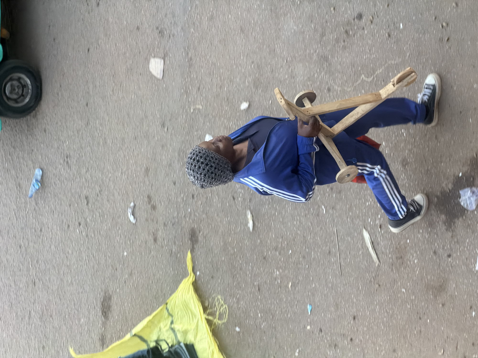 A woman is walking on a paved surface, carrying a wooden toy. She is wearing a gray knitted hat, a blue tracksuit with white stripes, and black sneakers. She is carrying a wooden toy scooter with two wheels. The ground is gray and appears to be asphalt, with some scattered debris. In the background, a yellow tarp is visible, along with the wheel of a vehicle. The scene is set in Bukuru, Nigeria.