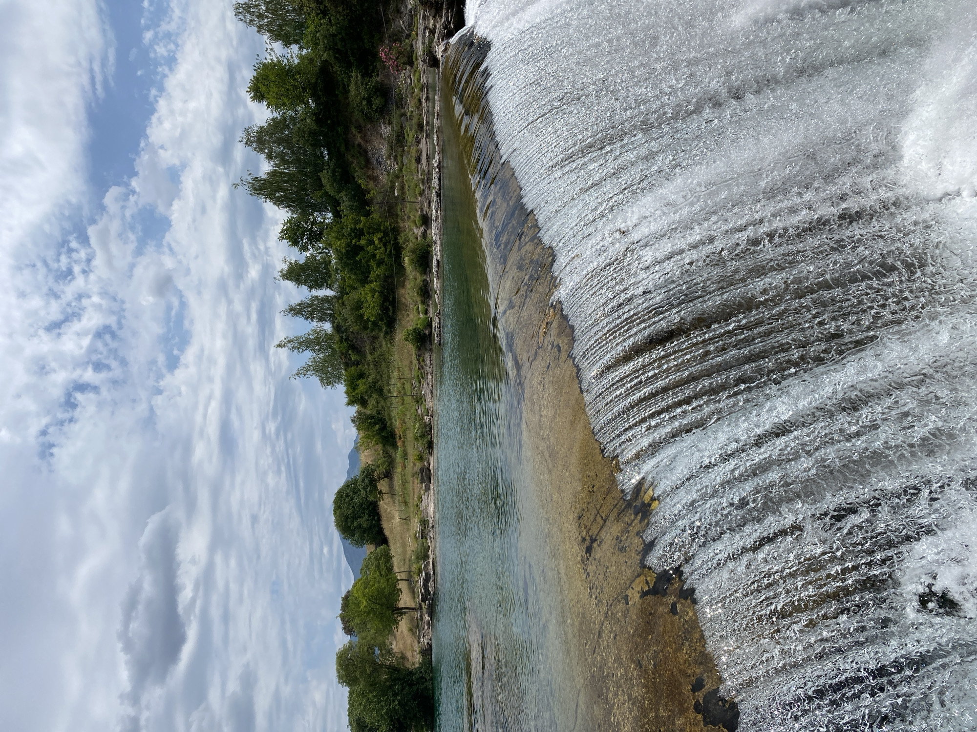 The image presents a serene natural landscape, captured horizontally, likely near Podgorica, Montenegro. It features a wide river flowing over a low weir or natural rock formation, creating a gentle, cascading waterfall under a partly cloudy sky.
In the foreground, the right side of the image is dominated by the dynamic movement of water forming a wide waterfall. The water tumbles down in numerous textured, frothy white cascades, glistening under the daylight. To the left of the waterfall, the river continues, appearing calm and remarkably clear, revealing a rocky, sandy riverbed below. The water in this section shows beautiful turquoise and green hues.
The left bank of the river is densely lined with lush, vibrant green trees and bushes, suggesting abundant vegetation. Beyond the immediate riverbank, the terrain rises gently, covered in lighter green grass and sparse trees. In the distance, faint outlines of mountains or hills are visible, blending with the horizon.
The sky above is bright blue, dotted with scattered white and soft grey cumulus clouds, indicating a fair-weather day, likely late morning or early afternoon. The overall atmosphere is one of tranquility and natural beauty. There are no people, visible objects of human intervention apart from the potential weir, or any discernible text in the frame.