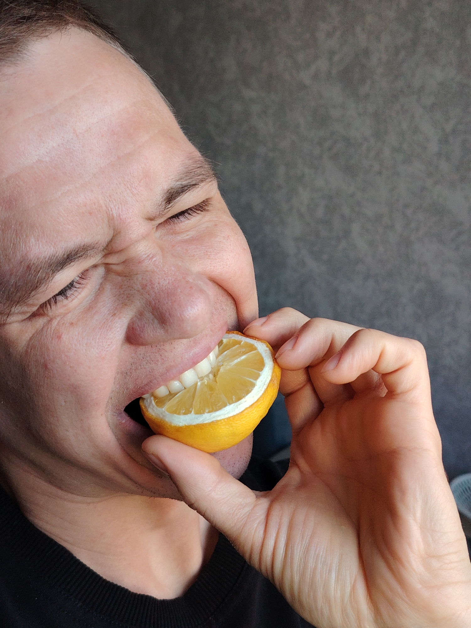 A close-up shot shows a person biting into a half-cut lemon, their face contorted in a grimace likely due to the sourness. The person's eyes are closed tightly, and the facial muscles are tense. The lemon is held in their left hand, positioned against their mouth. The background is a blurry gray. The location of the image cannot be determined from the content of the image.