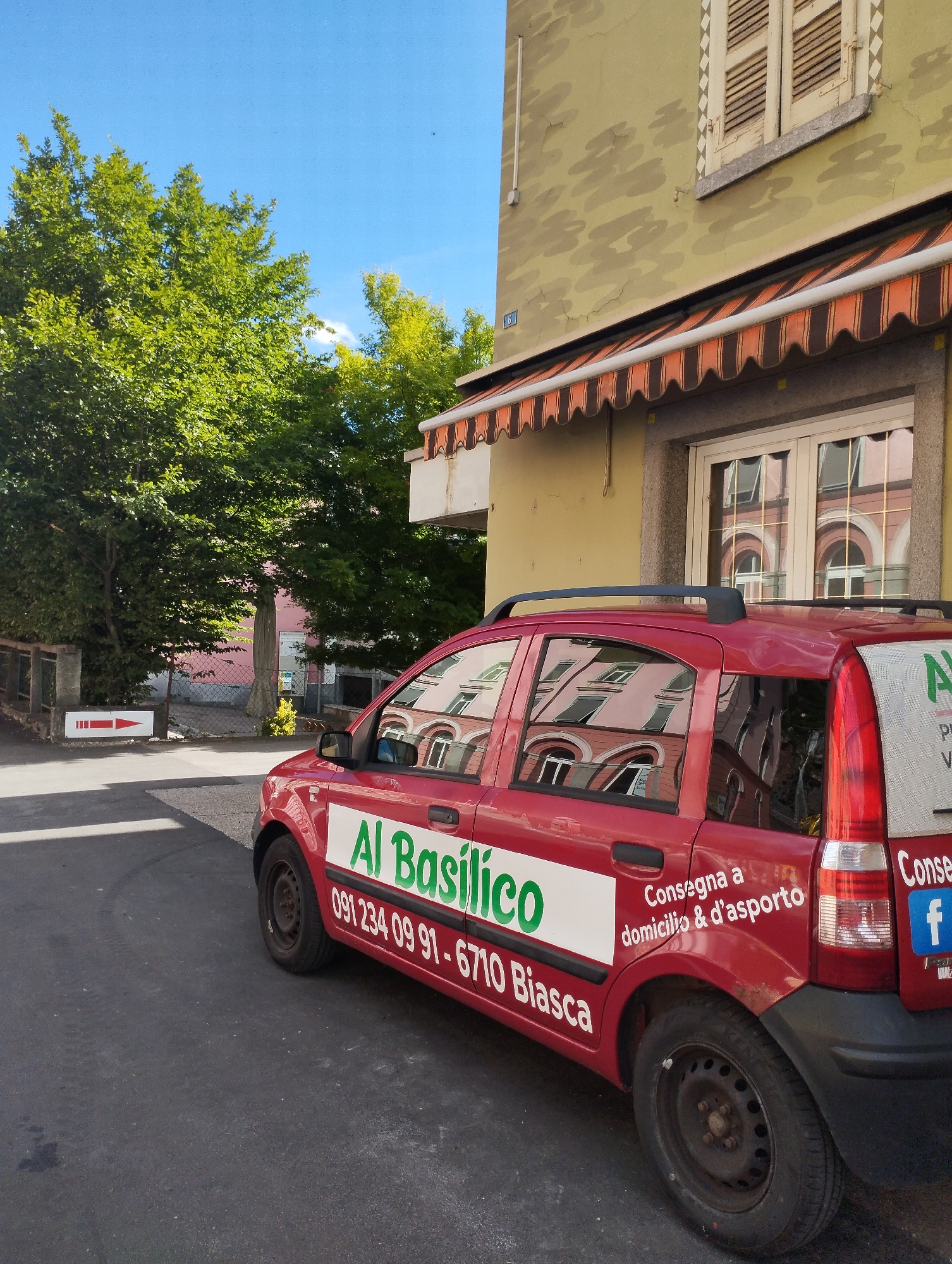 A red car with "Al Basilico" written in green on its side is parked on a street. The car has a delivery service advertisement on its rear quarter panel. The building behind the car has a yellow facade and a striped awning. Trees are visible on the left side of the image. A red arrow sign is also present on the left side of the street.