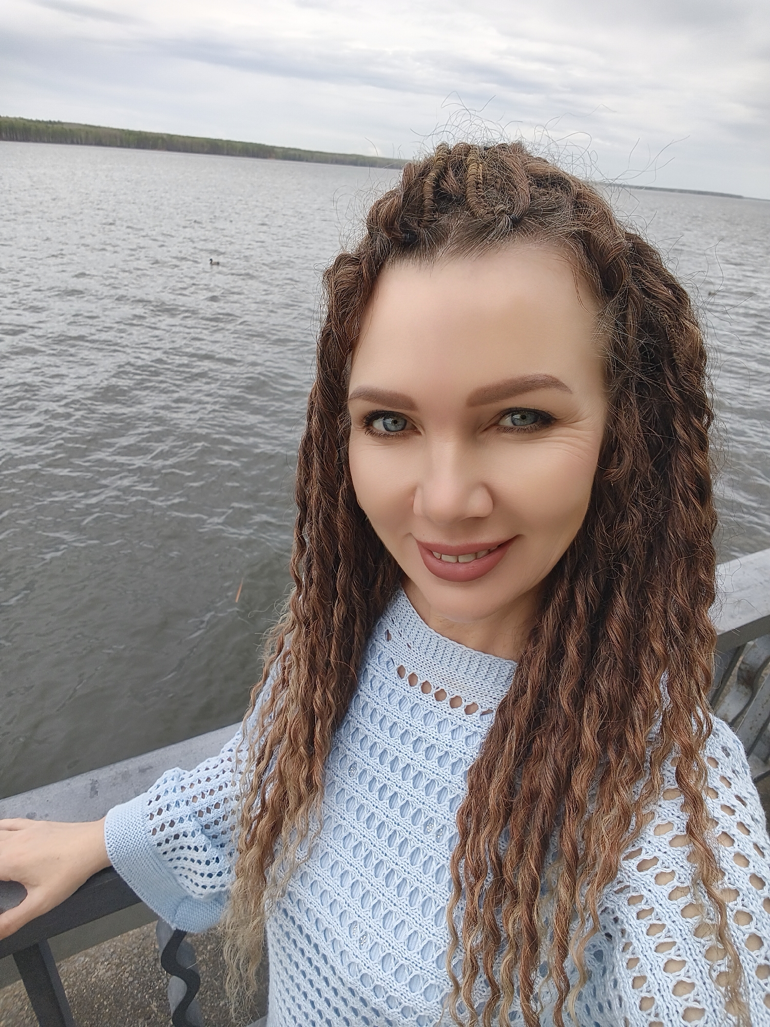A woman with light skin, braided brown hair, and blue eyes smiles while taking a selfie. She is wearing a light blue knitted top and is leaning against a gray metal railing. Behind her is a body of water, likely a lake or wide river, with a distant treeline visible on the opposite shore under an overcast sky. There is some evidence of a city in the background. The location appears to be a waterfront area or a bridge.