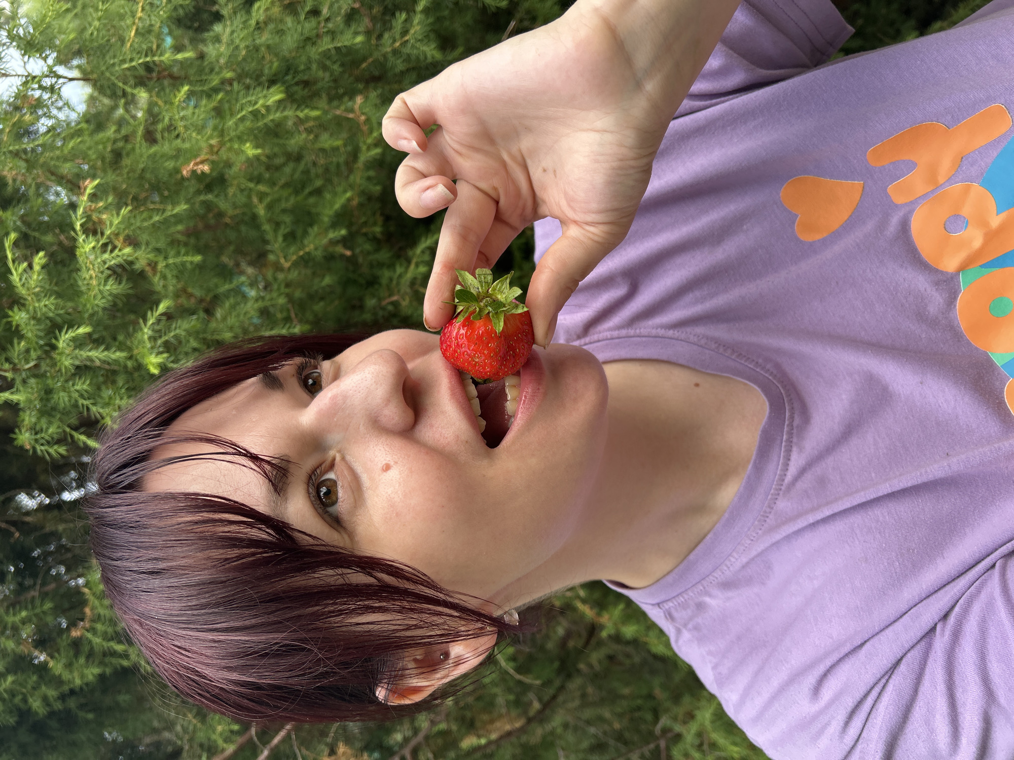 A woman with short, dark red hair is holding a strawberry up to her open mouth, about to eat it. She is wearing a light purple t-shirt with an orange heart and text on it. The background is out of focus and appears to be a lush green hedge or shrubbery, suggesting an outdoor setting. The image was taken in the city of Фоки, Russia.