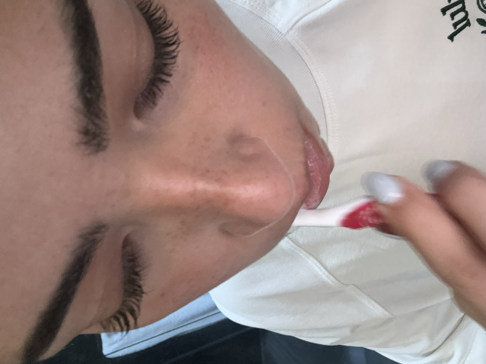A person with dark eyebrows and long eyelashes is brushing their teeth. The toothbrush is red and white. The person is wearing a white shirt with green writing. The setting appears to be indoors. The location is unknown and is not identifiable from the provided information.