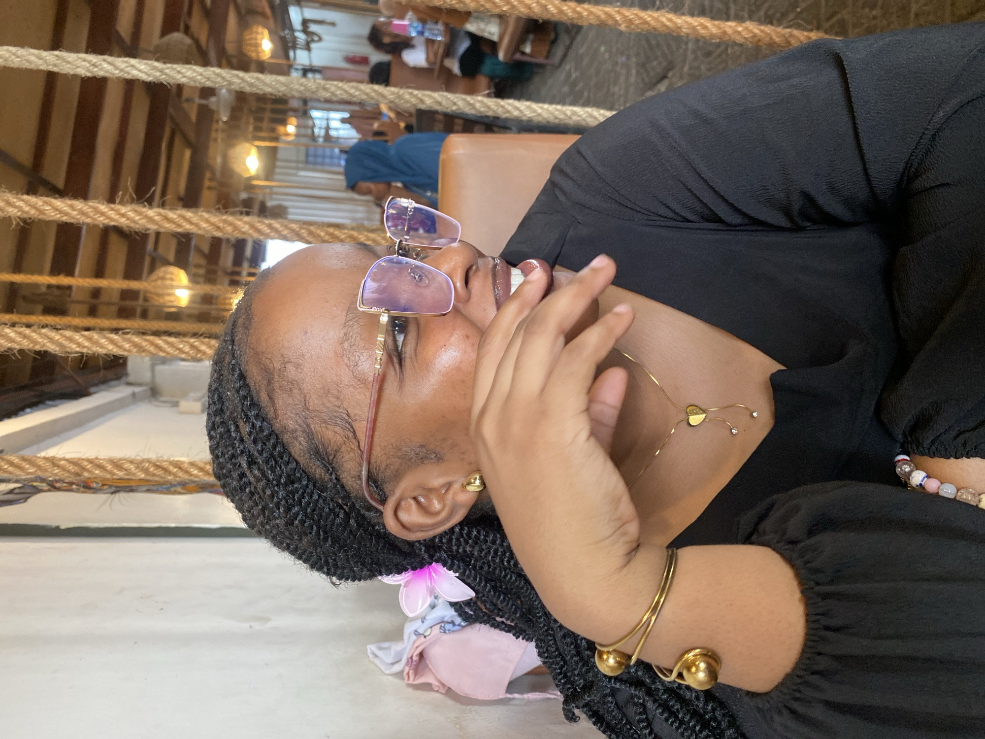 A person is angled in the foreground, positioned against a backdrop of indoor and outdoor elements. The person is wearing black clothing, glasses with light-colored frames, and jewelry, including a necklace, earrings, and bracelets. The person's hair is styled in braids.
Behind the person is a seating area with furniture. Ropes and wooden beams create a decorative element in the background, while hanging lights provide illumination. Other patrons sit at tables in the background. The scene suggests an establishment in Dar es-Salaam, Tanzania.