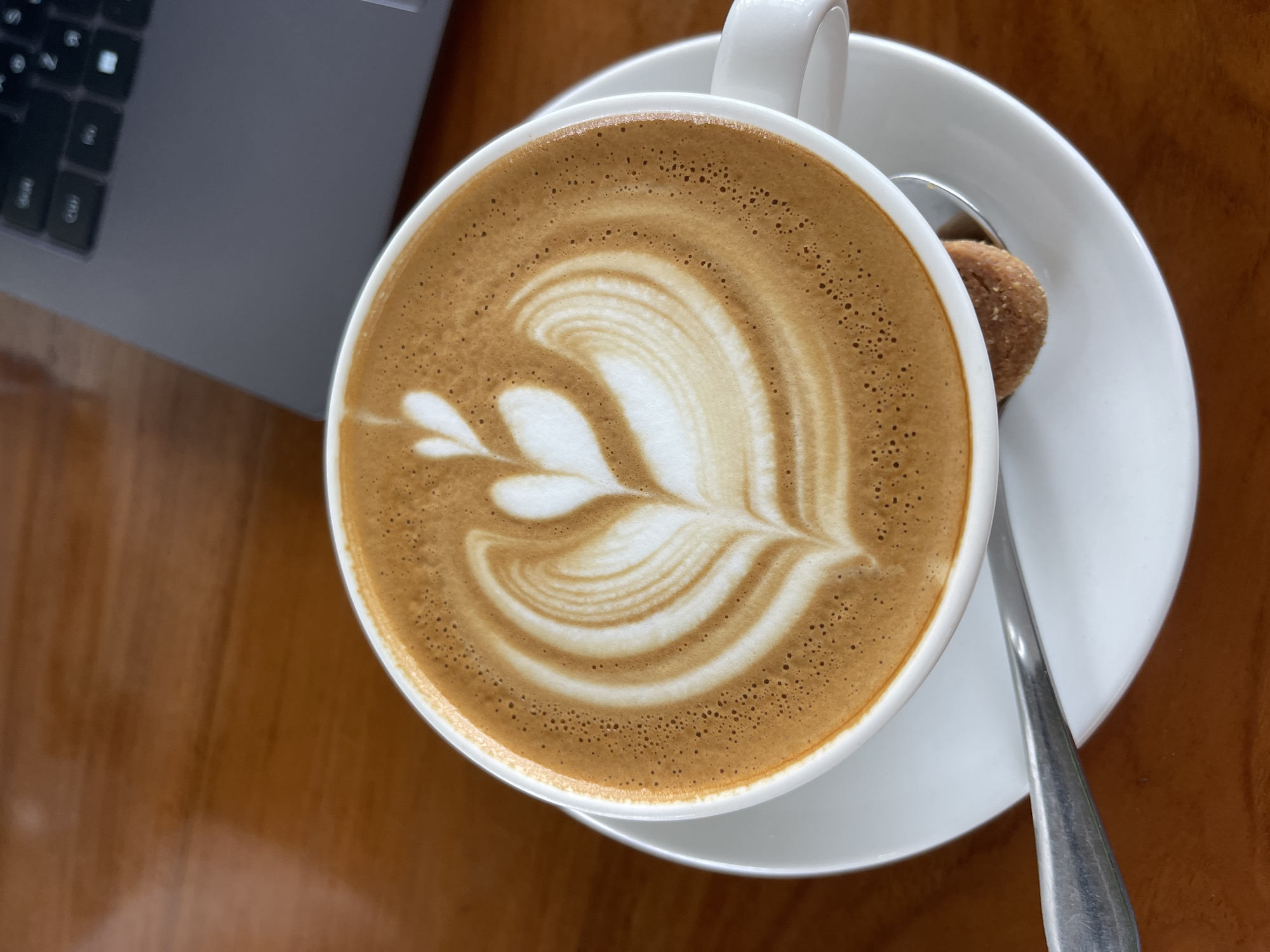 An overhead, close-up shot captures a white mug of coffee with latte art, presented on a white saucer. The latte art forms a symmetrical pattern resembling a heart with wings. A small, circular cookie rests on the saucer, partially submerged by a silver spoon. The mug and saucer sit on a polished wooden surface. In the upper right corner of the frame, a portion of a gray laptop is visible. The scene is devoid of people, and the lighting appears natural, possibly from a nearby window. The setting is likely a cafe or a workspace, given the presence of the laptop and coffee. This image may have been taken in North Kuta, Indonesia, a region known for its cafes and vibrant atmosphere, though there is no direct visual evidence to confirm this location.