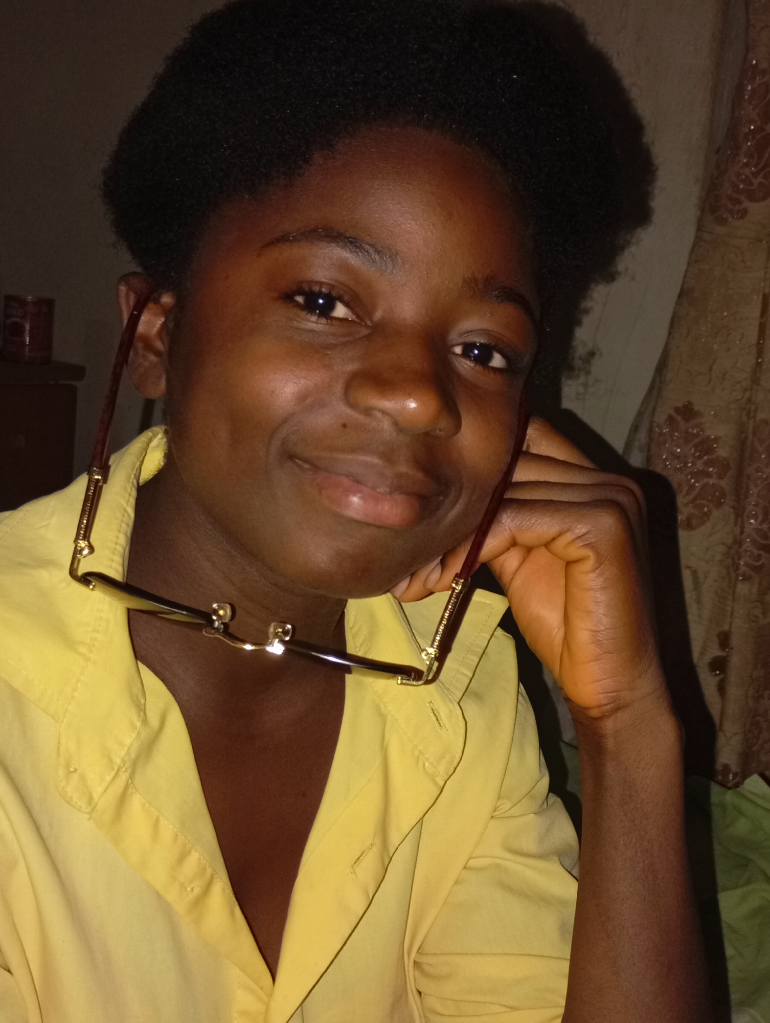 A close-up shot depicts a Black person wearing a yellow collared shirt. They have dark hair, dark eyes, and are smiling slightly. The person is holding a pair of glasses with a brown frame in one hand, the frame resting on their chest. The person's hand is resting on the face. The background includes a curtain with a patterned design. Jos, Nigeria is not evident in the image. No specific location in the city of Jos can be identified.