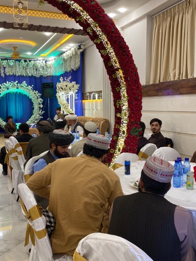 The image depicts a group of men gathered inside a decorated event space, likely in Kandahar, Afghanistan. The attendees are primarily dressed in traditional attire, with some wearing turbans or distinct caps. They are seated at tables covered with white cloths, where bottled water and other items are placed. The room's decoration includes a large, curved floral arrangement of red and white flowers, along with additional decorative elements like floral wreaths and blue backdrops. The setting suggests a formal event, possibly a wedding or a traditional gathering.