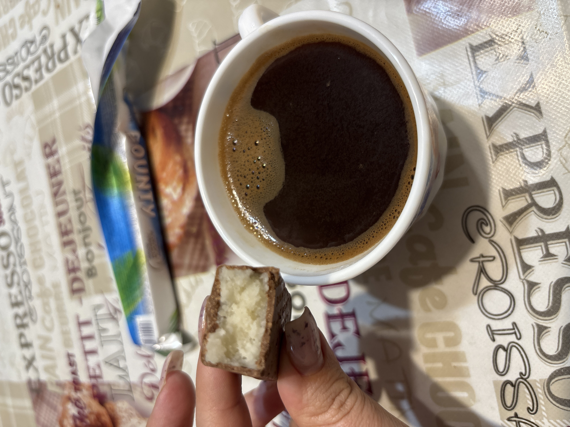 A hand holding a broken chocolate bar is positioned over a white mug of black coffee. The chocolate bar has a white filling. The mug and the chocolate bar rest on a patterned surface with text in French. The pattern includes words like "EXPRESSO," "CROISSANT," and "CHOCOLAT." The scene is brightly lit, suggesting it is indoors during the daytime. There is no information available about the location in the city of Zubov, Russia.