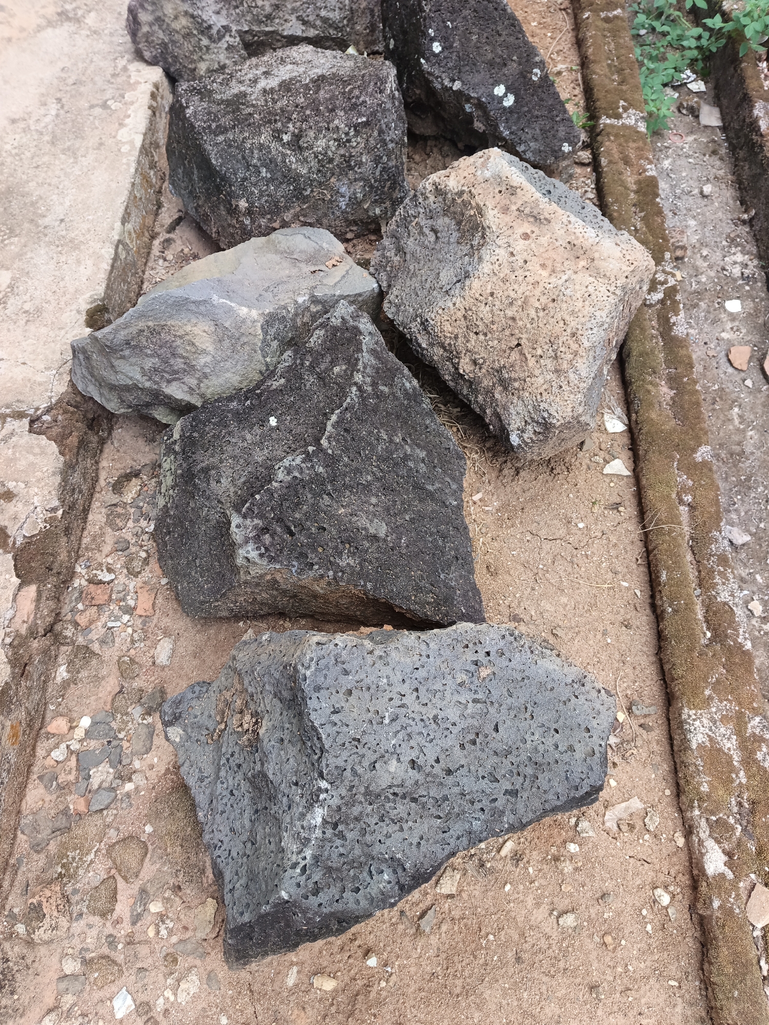 The image shows a pile of large, irregular rocks of varying colors and textures scattered on the ground. The rocks appear to be natural, with some exhibiting rough, porous surfaces and others smooth, layered surfaces. The ground beneath them is composed of dirt and small pebbles, with some dried vegetation. To the right of the rocks, a trench or channel is visible, lined with moss-covered stones. Some broken pieces of terracotta or pottery are also scattered in this area, suggesting a possible archaeological or historical site. The overall setting appears to be outdoors, possibly in a rural or undeveloped area. The specific location in Terbanggi Marga, Indonesia, is not visually evident from the image itself, but the context suggests it could be an archaeological excavation or a historical preservation site. There are no people visible in the image.