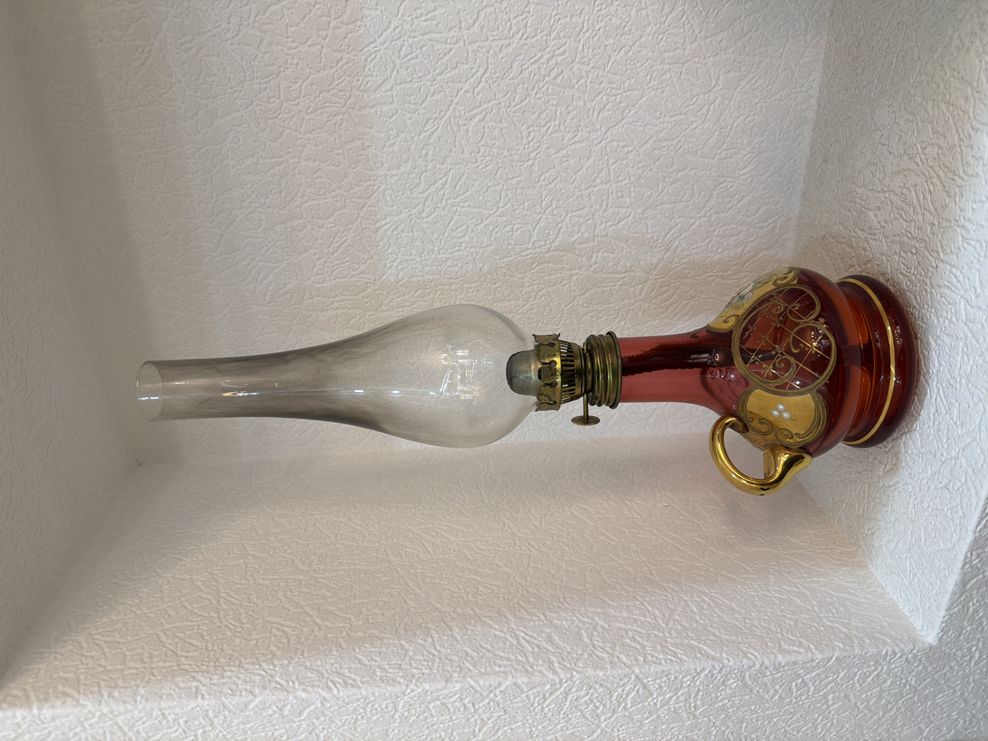 A glass oil lamp with a red glass base, adorned with gold decorative accents, sits on a white shelf in a corner. The lamp has a brass wick holder and a clear, elongated glass chimney. The background is a white, textured wall. The scene is indoors, likely a home. There are no people present in the image. The location of this image is not Sabirabad, Azerbaijan.