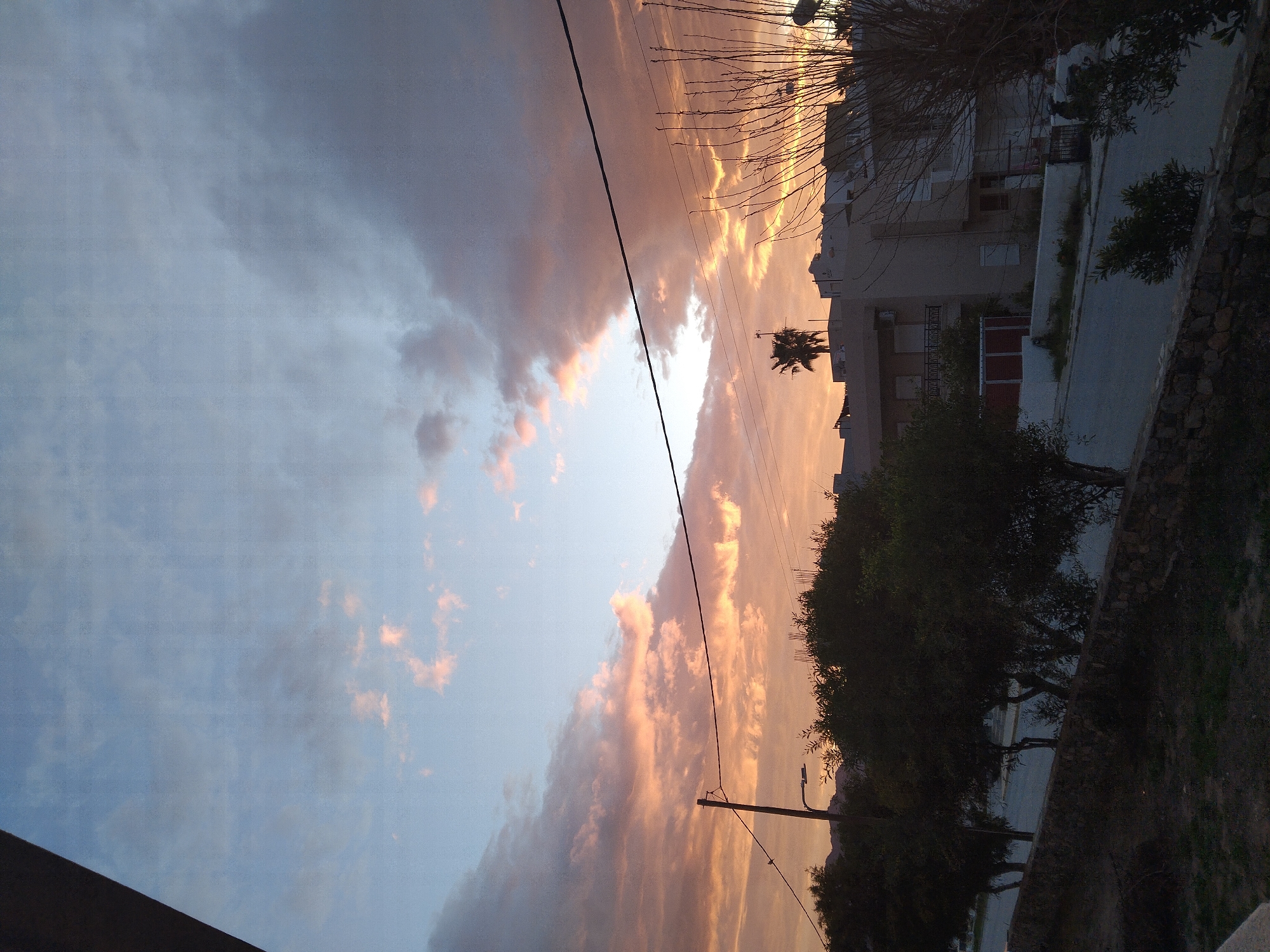 The image shows an evening scene in Καρδάμαινα, Greece. The sky is split between a light blue and a dramatic orange, possibly indicating a sunset or sunrise. Wisps of clouds are present in both the blue and orange areas. A thick black cable runs vertically through the center of the image. On the right side are buildings, foliage, and trees. A palm tree is visible, standing near a building. A streetlight is to the right of the frame. The photo is taken from an elevated position, looking downwards towards the landscape.