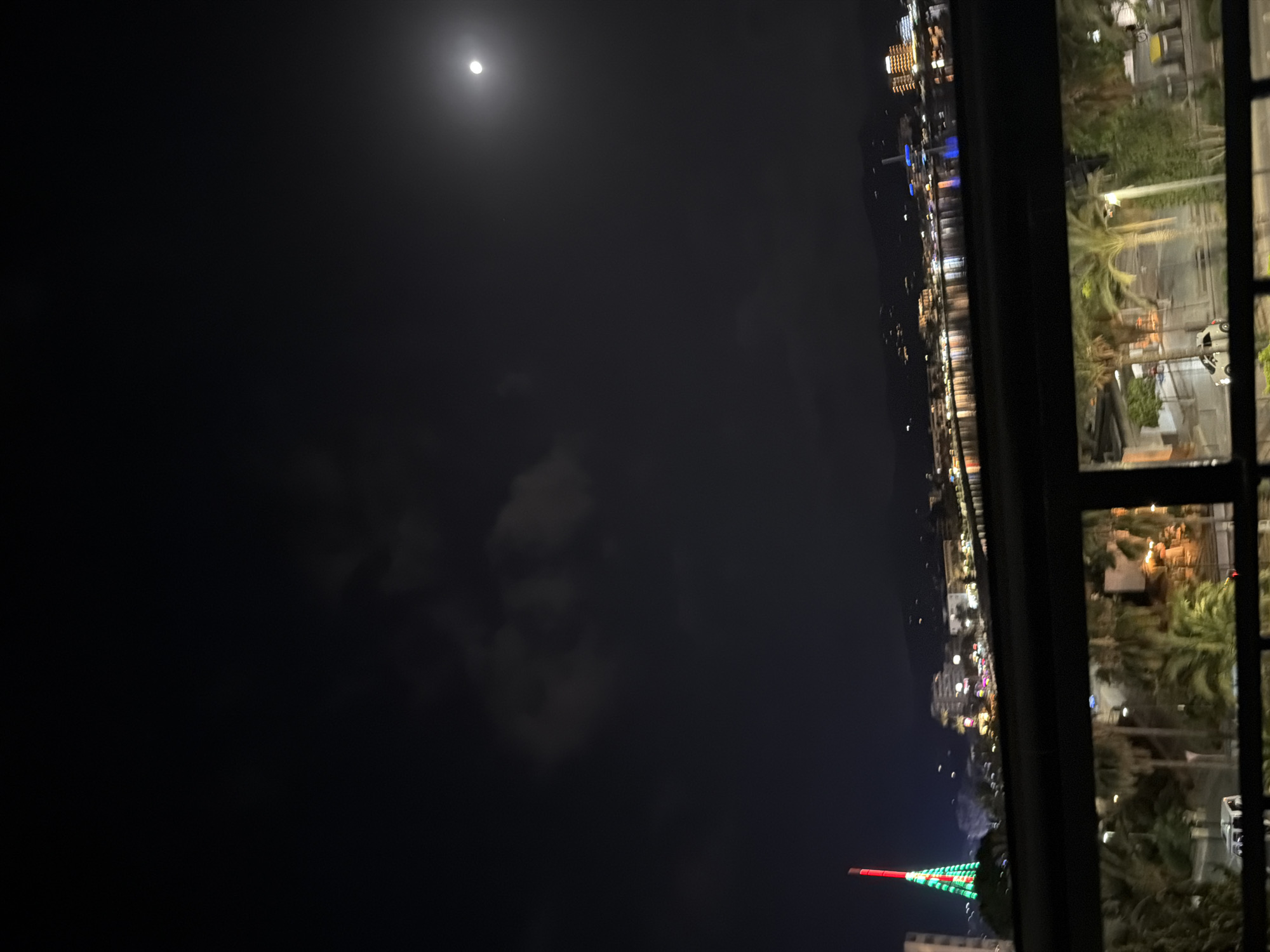 The image captures a nighttime view of the bay in Sant Antoni De Portmany, Spain, as seen from an elevated position. A bright full moon is visible in the dark sky, partially obscured by wispy clouds. In the distance, the city skyline is illuminated with numerous lights reflecting on the calm water of the bay. A prominent, illuminated tower, possibly a landmark or decorative structure, stands out on the left side of the cityscape, adorned with green and red lights. The foreground features a dark, horizontal railing, suggesting the viewpoint is from a balcony or a similar structure. Below the railing, palm trees and some buildings are visible, indicating a coastal urban environment. There are no discernible people or specific actions taking place in the image.