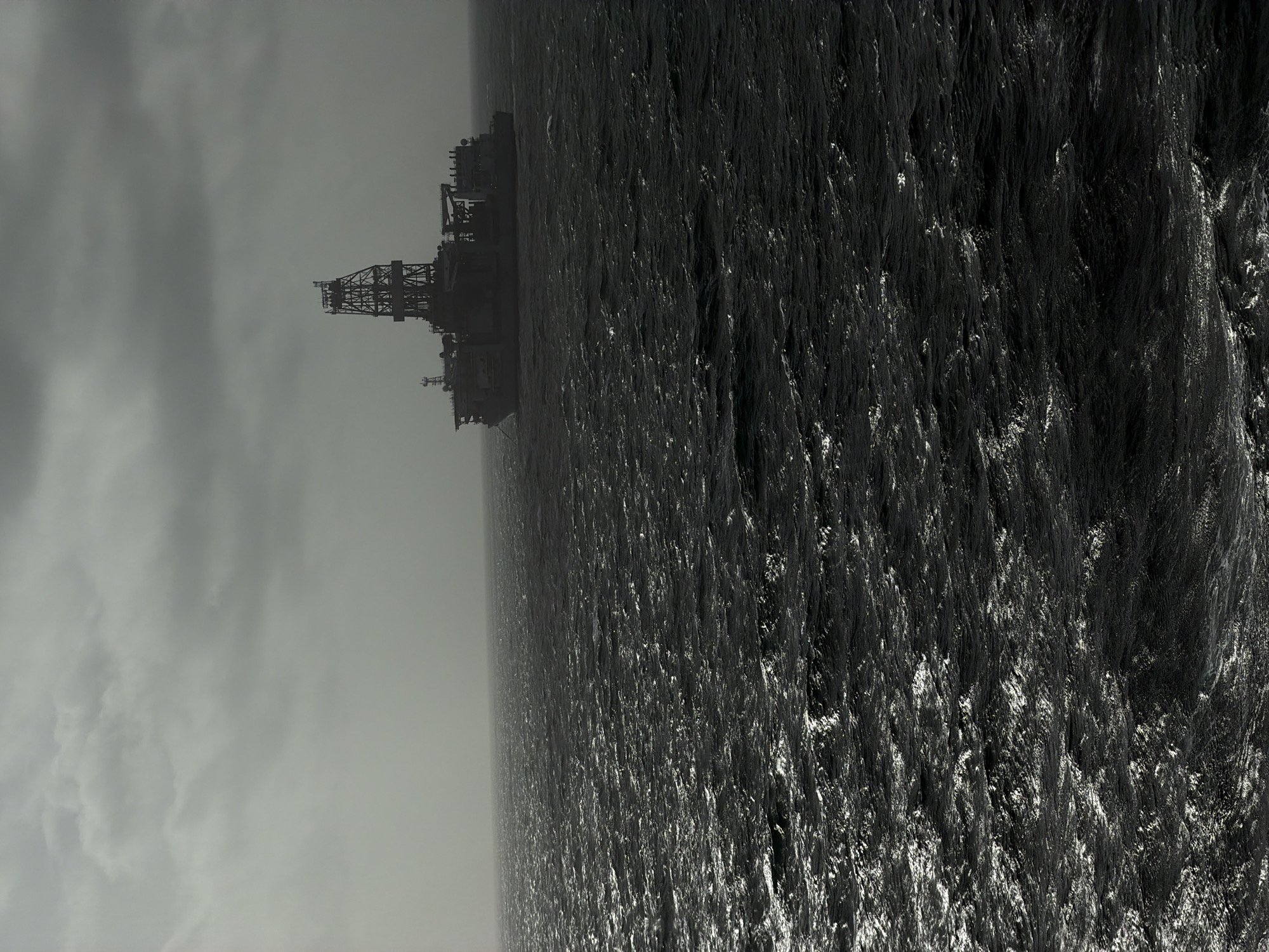 The image depicts an offshore oil platform surrounded by rough, dark water under a cloudy, gray sky. The platform is located in Unknown, Unknown. The structure of the platform is complex and appears to be made of metal. The water's surface is highly textured and reflective, indicating rough sea conditions.