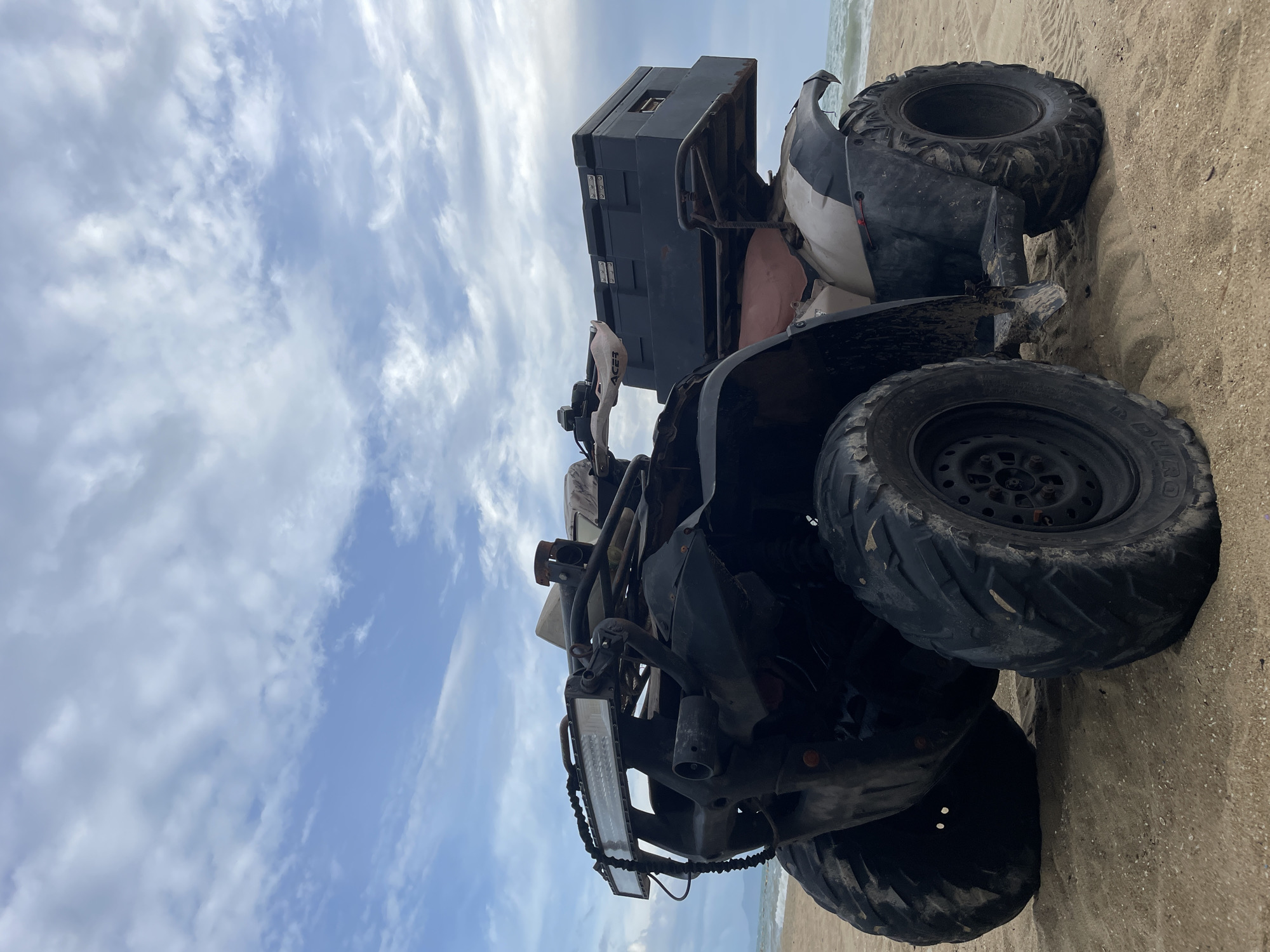 An all-terrain vehicle (ATV) is parked on a sandy beach with the ocean and a cloudy sky in the background. The ATV is black and beige with large, knobby tires. It has a prominent LED light bar mounted on the front. A black storage box is attached to the rear rack. The ATV appears to be positioned at a slight angle, facing towards the left of the frame. The sand is lightly rippled, suggesting it may be a tidal area. The water of the ocean is visible on the right, with small waves. The sky is filled with scattered clouds, some appearing white and wispy, others darker and more dense, indicating a partly cloudy day. The scene is likely located on one of the beaches in or around Pattaya City, Thailand, given the typical climate and coastal landscape. No people are visible in the image.