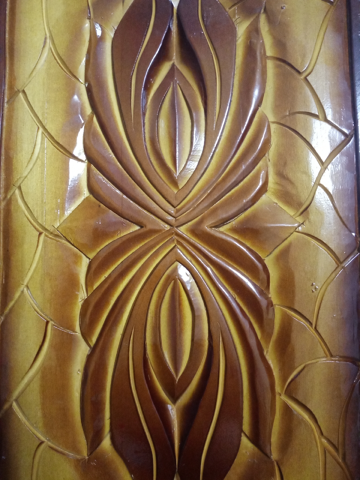 The image displays a close-up view of a richly carved wooden panel, presenting a symmetrical and intricate design with a glossy, varnished finish. The material appears to be wood, characterized by warm tones of brown and yellowish-brown, with variations in shade that highlight the three-dimensional carving.
The central feature of the panel is an elaborate, vertically aligned, bilaterally symmetrical motif. This main design is composed of multiple layers of smoothly curved, raised forms, resembling stylized plant elements like intertwined leaves, petals, or abstract floral patterns. These elements meet at a central point, creating a cohesive, organic flow. The glossy surface reflects ambient light, accentuating the contours and depths of the carving, and emphasizing its relief texture.
Flanking the central design, and extending towards the edges of the frame, is a repeating background pattern of interlocking curved lines. This pattern creates a textured, scale-like or quilted effect, offering a contrasting, more geometric yet still organic, backdrop to the fluid central motif. The interplay of light and shadow across both the main and background carvings underscores the craftsmanship and intricate detailing of the piece.
No people are visible in the image, nor is there any discernible text. The scene depicts a static, decorative object, likely a component of furniture, a door, or architectural ornamentation, reflecting traditional or artisan woodworking. The image provides no specific time of day or weather cues, as it focuses solely on the indoor object.