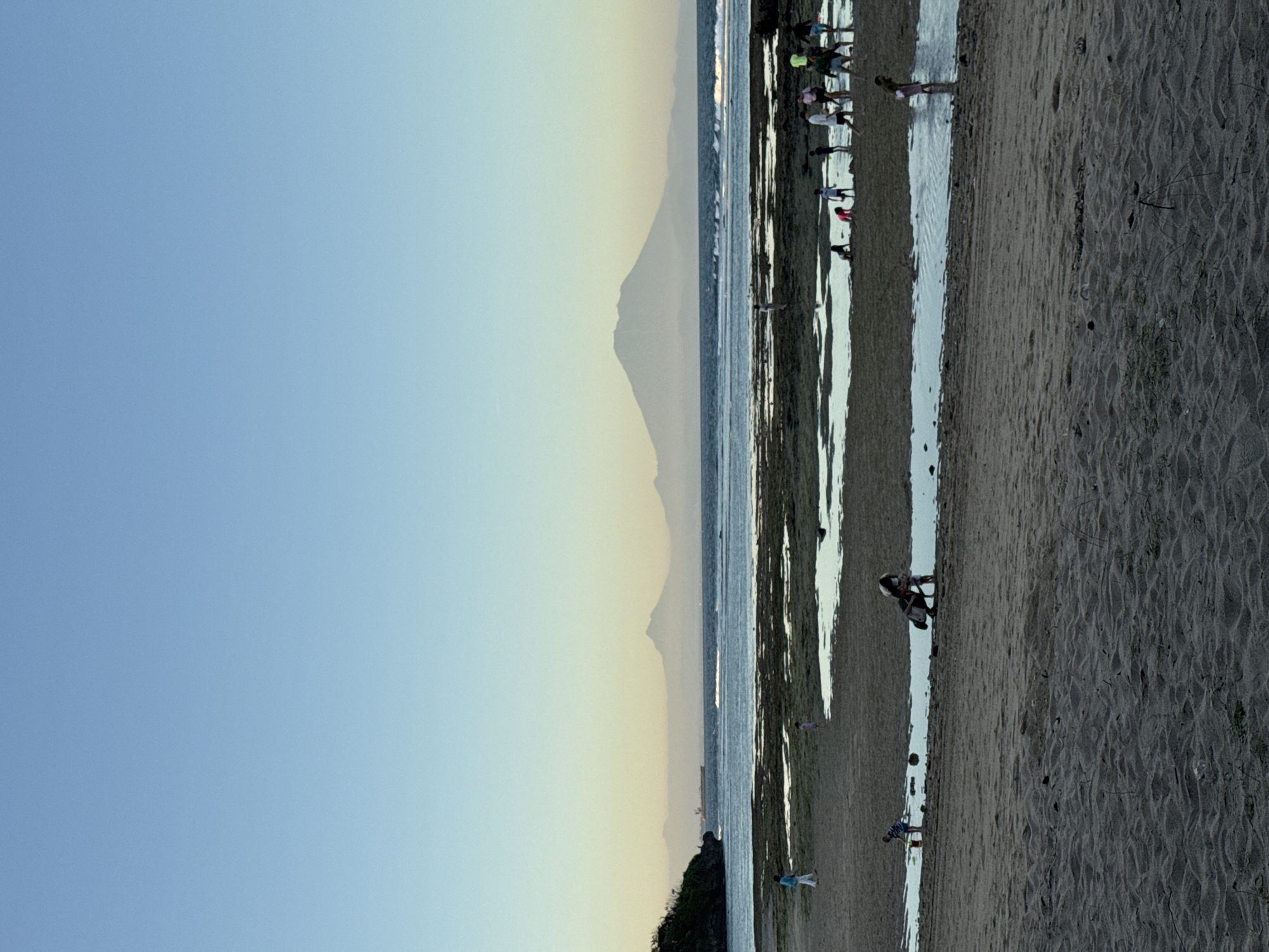 The image shows a beach landscape at sunset in Kuta Selatan, Indonesia, with a mountain visible in the background. A group of people are standing in the shallow water, possibly clamming or playing. The beach is sandy with some vegetation near the edge. The sky is clear with hues of blue and yellow indicating the setting sun.