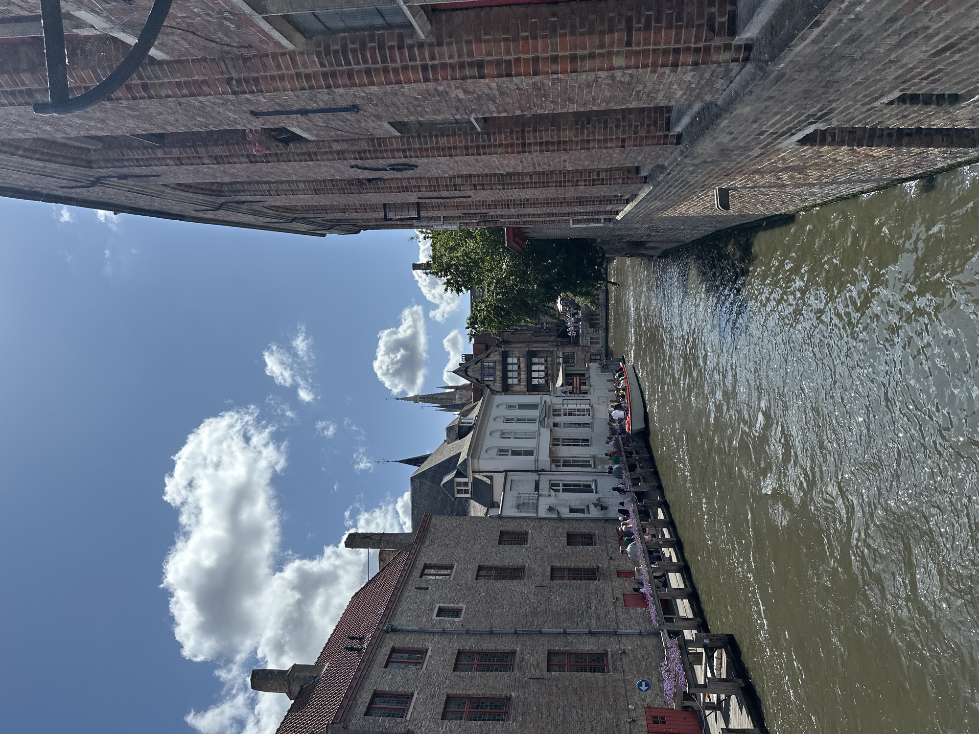 This image captures a charming canal scene in Bruges, Belgium. The canal, with its muddy, rippling water, reflects the bright blue sky and fluffy white clouds. Along one side of the canal, a wooden boardwalk is lined with people, suggesting a popular tourist spot. Several boats are docked or moving along the canal, with one boat in particular crowded with passengers, hinting at a boat tour.
The architecture is characteristic of Bruges, featuring historic brick buildings and houses with distinctive gabled roofs and multiple windows. Some buildings are made of weathered brick, while others are painted white or light grey. The juxtaposition of old and new is evident, with some structures appearing well-maintained and others showing signs of age. Trees with lush green foliage provide a natural contrast to the stone and brick structures.
The time of day appears to be daytime, likely late morning or early afternoon, given the strong sunlight and clear sky. The weather is pleasant, with sunshine and scattered clouds. The overall mood is serene and picturesque, evoking a sense of historical charm and leisure. The presence of numerous people enjoying the scenery suggests a lively yet relaxed atmosphere. There is a small blue arrow sign visible on the boardwalk, likely indicating directions.