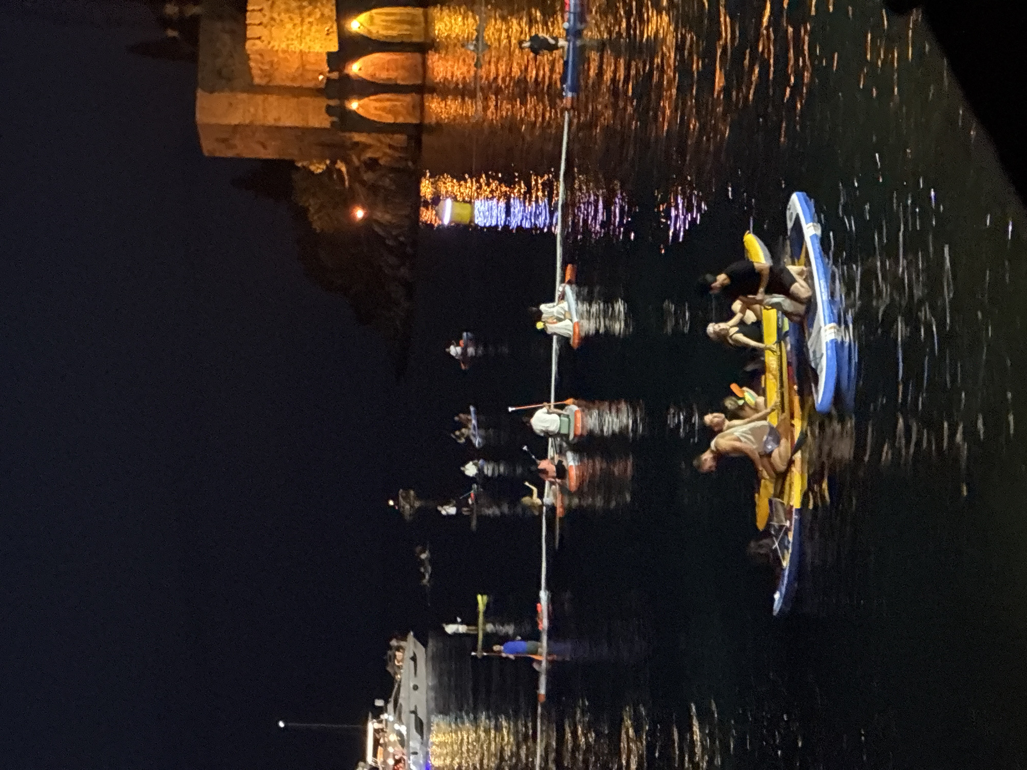 The image depicts a nighttime scene on the water, likely in Alanya, Turkey, given the architectural style of the illuminated structure in the background. The main subjects are groups of people on paddleboards and kayaks. The individuals are engaged in what appears to be a leisurely activity on the water, possibly a guided tour or a nighttime paddle event.
In the foreground, several paddleboards and kayaks are clustered together. Some people are standing on the paddleboards, while others are seated or lying down. The individuals are dressed casually, and some appear to be interacting with each other. One paddleboard has a blue and white design, and another is predominantly yellow.
In the background, a stone structure, possibly a fort or a historical building, is illuminated with warm, golden lights. The lights are reflected on the dark water, creating a shimmering effect. Further in the background, more lights, some appearing blue, are visible, suggesting a city or a waterfront promenade. The overall atmosphere is serene and tranquil, with the dark sky and the illuminated reflections contributing to a peaceful ambiance.
The time of day is clearly night, indicated by the darkness of the sky and the artificial lighting. The weather appears calm, as the water is relatively still, with only minor ripples. No text is visible in the image. The scene evokes a sense of enjoyment and relaxation as people partake in a water activity under the night sky.