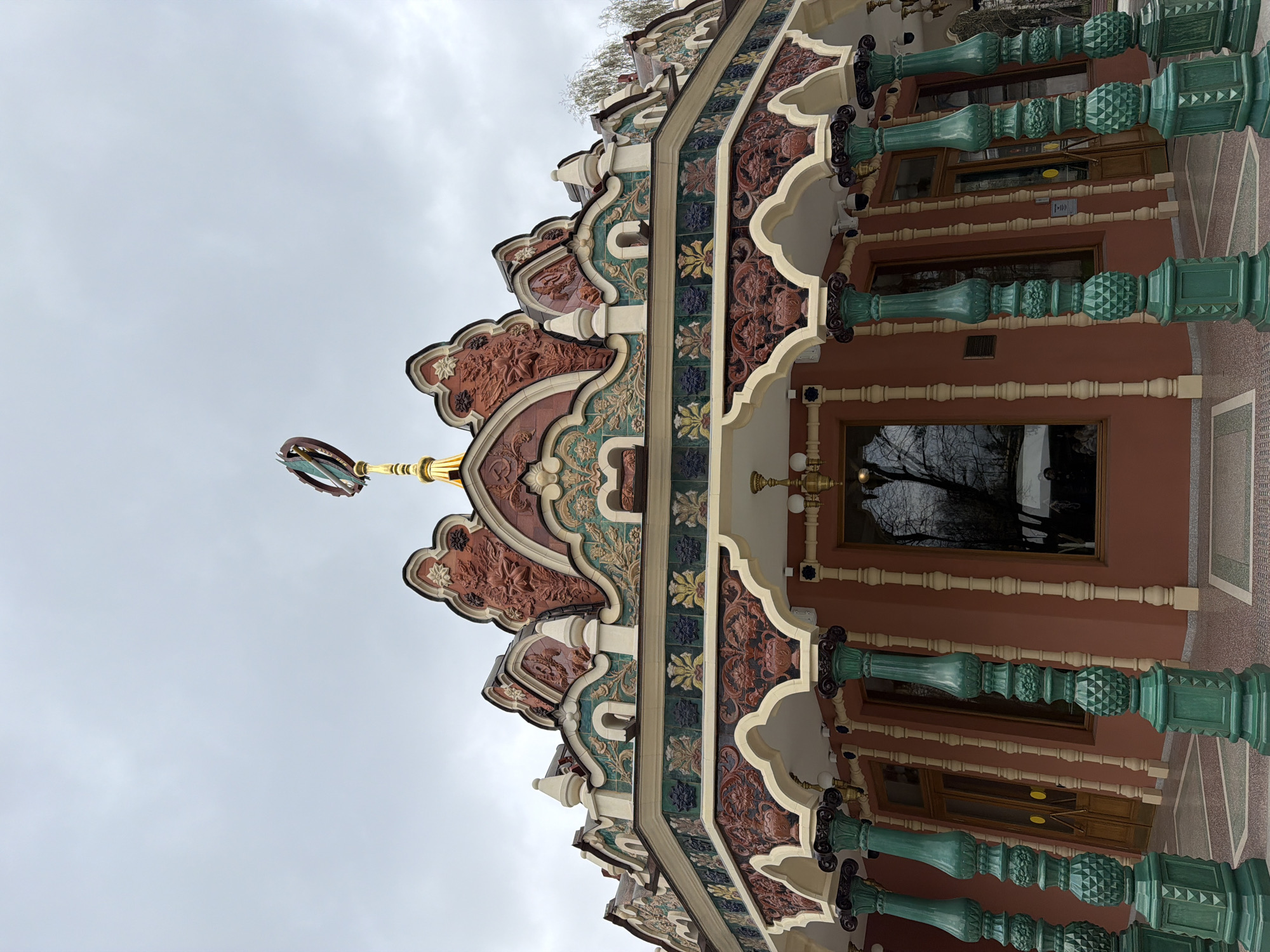The image depicts an ornate building facade in Moscow, Russia. The building features elaborate decorative tiling in shades of red, green, and cream, as well as green pillars. The roofline has a complex, multi-tiered design. Atop the building sits a golden spire with an emblem attached. The sky is overcast. The photograph appears to be taken from a low angle, looking up at the building. The image is rotated 90 degrees.
