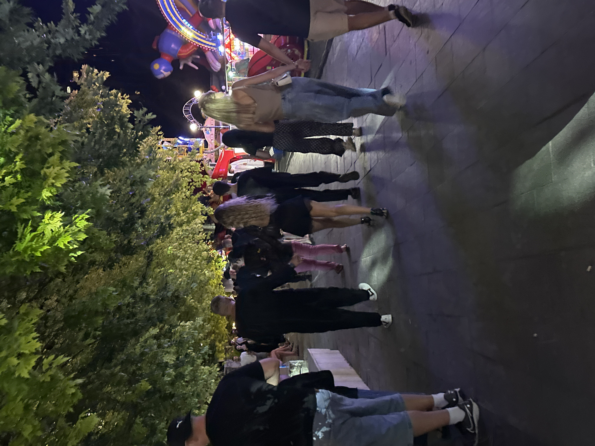 The image depicts a nighttime scene at an amusement park or fairground. Several people are walking on a paved walkway lined with trees. In the background, amusement rides with colorful lights are visible. The atmosphere appears festive and crowded. It is not possible to determine the specific location within the city of Odesa, Ukraine, or any other city from the provided image. The people are dressed in casual attire.