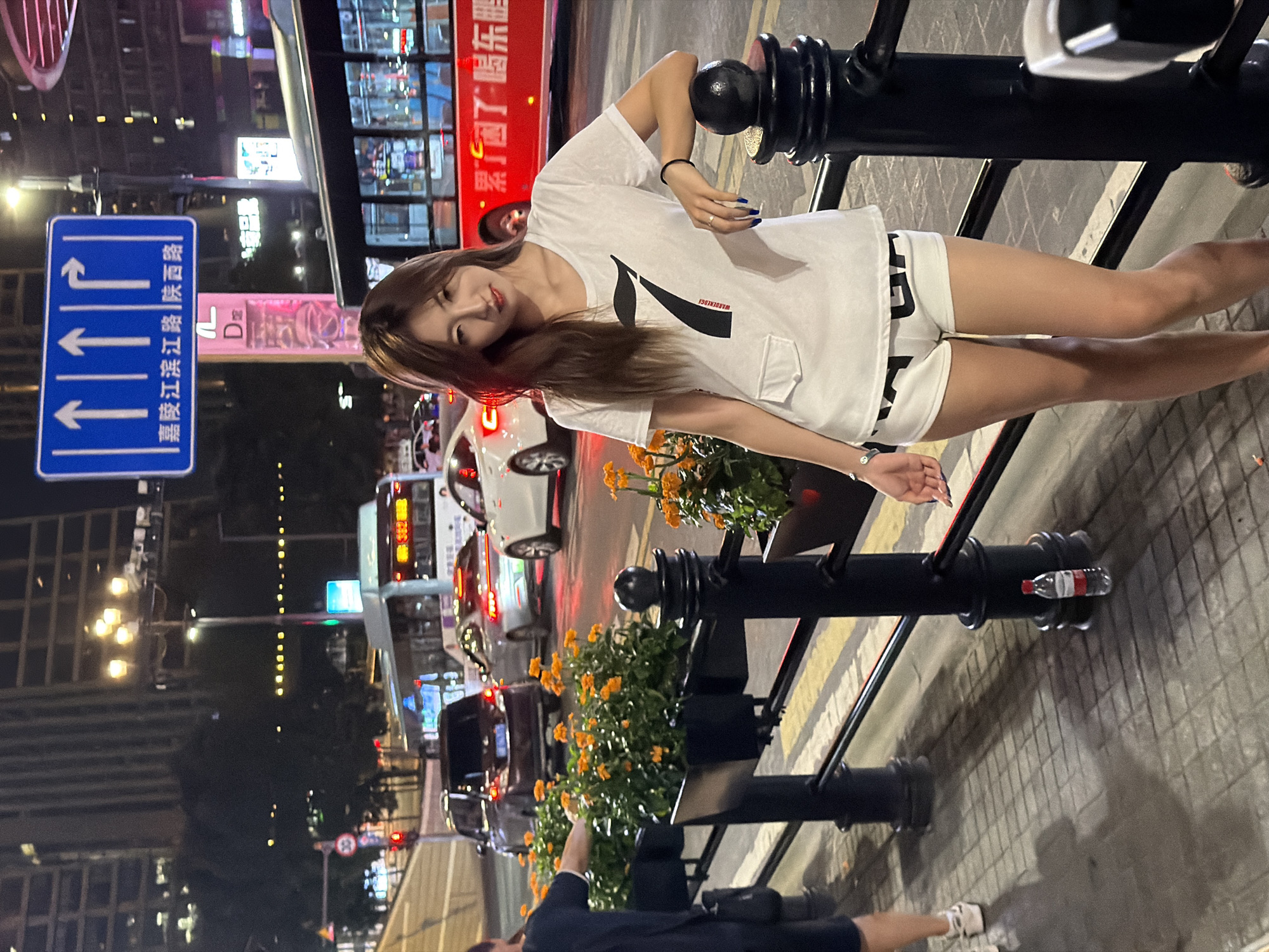 A young woman with long brown hair stands on a sidewalk at night in Yuzhong District, China. She is wearing a white t-shirt with a large black "7" graphic and white shorts. She is leaning against a black metal railing. In the background, there is a busy street with traffic, including cars, a bus, and a traffic signal. The sidewalk is paved with cobblestones, and there are planters with orange flowers along the railing. A bottle of water is on the ground near the railing.