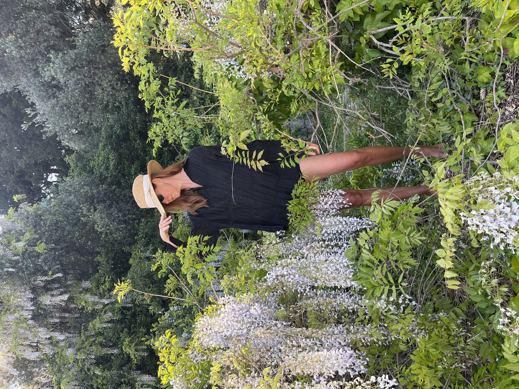 A woman is depicted amidst a lush outdoor setting, surrounded by abundant green foliage and blooming wisteria. She has long, reddish-brown hair and wears a straw hat with a white band, a black long-sleeved dress or tunic with a textured, possibly pleated fabric, and what appear to be tan or brown high-heeled boots. Her head is tilted, and her face is partially obscured by the hat and her hair, giving her a contemplative or serene appearance as she looks downwards or away.
The foreground and midground are dominated by dense wisteria vines, heavily laden with clusters of delicate light purple and white flowers. These flowers create a soft, ethereal border around the woman. In the background, various types of green trees form a rich, verdant backdrop, suggesting a natural garden or park environment.
The lighting indicates late afternoon or early evening, with a soft, warm glow illuminating parts of the foliage, suggesting a sunny day. The overall atmosphere is tranquil and natural. The woman appears to be posing for a photograph within this beautiful, floral landscape. The scene is set in Sveti Stefan, Montenegro. There is no visible text in the image.