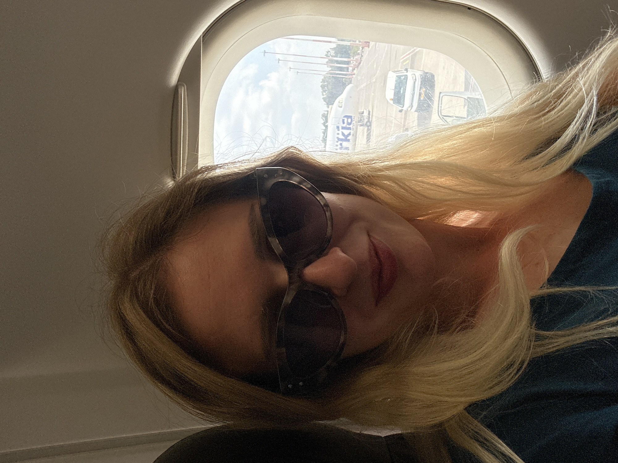 A woman with blonde hair, wearing sunglasses and a dark teal shirt, is seated on an airplane, taking a selfie by a window. Outside the window, an Arkia airplane is parked on the tarmac at Ben Gurion Airport, located in Yehud-Monosson, Israel. The sky is visible and appears to be partly cloudy. Several lights or poles and a white van are also visible on the airport grounds.
