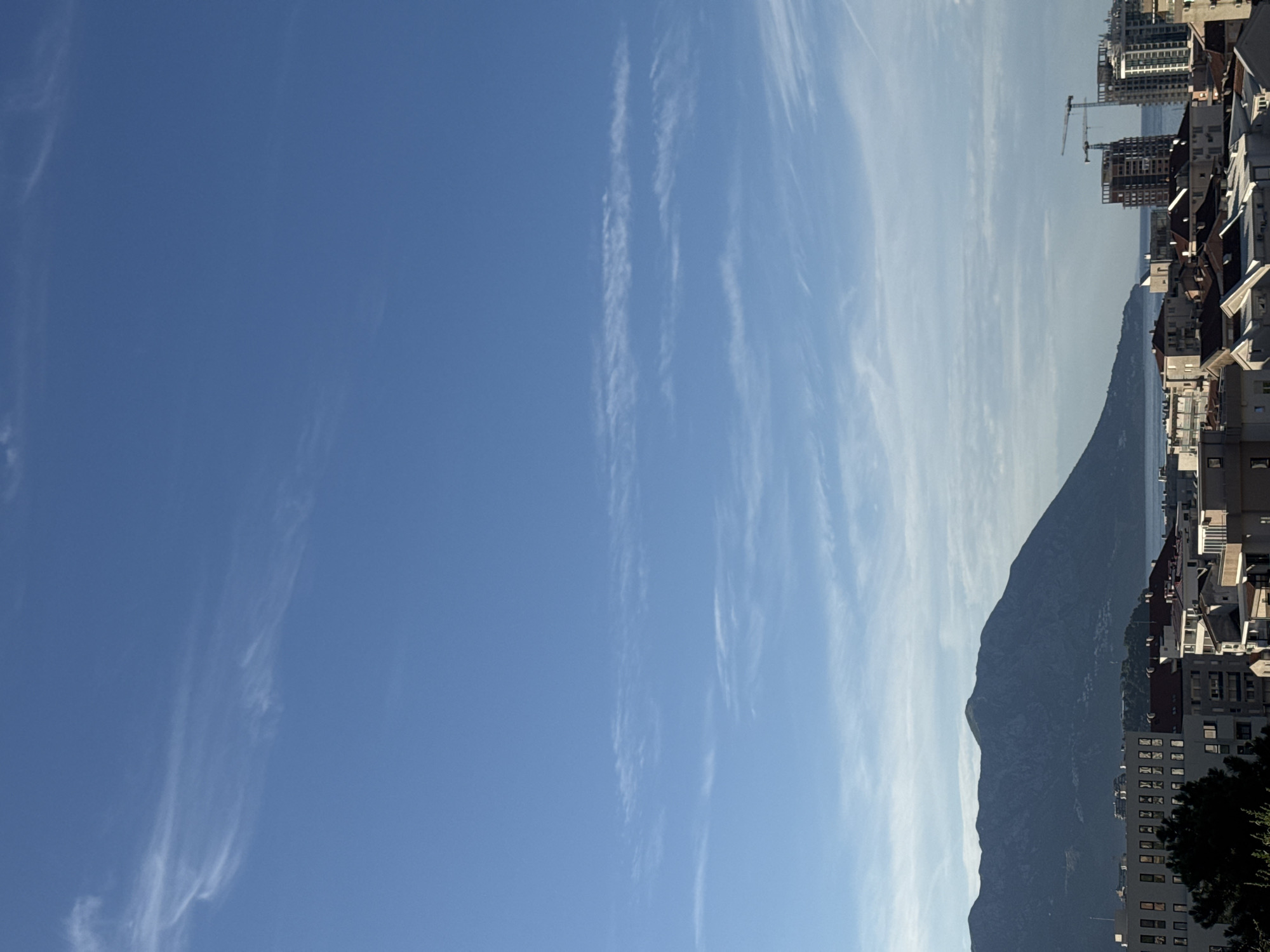 This is a wide shot of the city of Budva, Montenegro, taken from a high vantage point. The sky is a clear, bright blue with thin, wispy white clouds scattered across it. In the background, a large, dark mountain range dominates the horizon, its slopes covered in lush greenery. Below the mountains, the coastline stretches out, dotted with buildings and some construction sites. The foreground is filled with a dense cluster of buildings, varying in height and architectural style, with tiled roofs and multiple windows. Some trees are visible, adding a touch of nature to the urban landscape. The overall impression is that of a sunny day in a coastal city, with a mix of natural beauty and urban development.