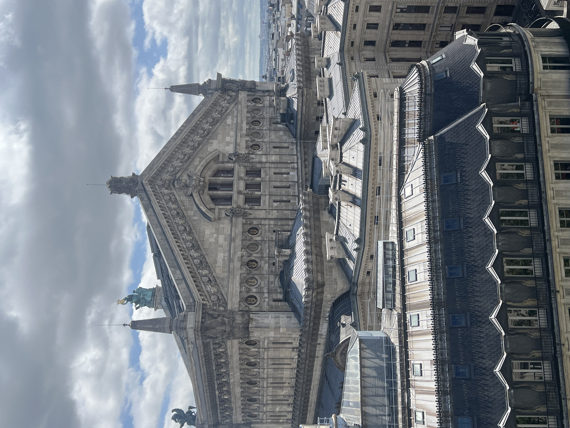 An aerial view captures the Palais Garnier, a grand opera house in Paris, France, on a cloudy day. The ornate stone building features a mansard roof with multiple dormer windows. To the left of the main structure, a bronze sculpture of a winged horse is visible on a pedestal. The opera house is surrounded by a dense urban landscape of similar buildings, also topped with mansard roofs and dormer windows, extending towards the horizon. The sky is filled with a mix of dark and light clouds, with patches of blue sky peeking through.