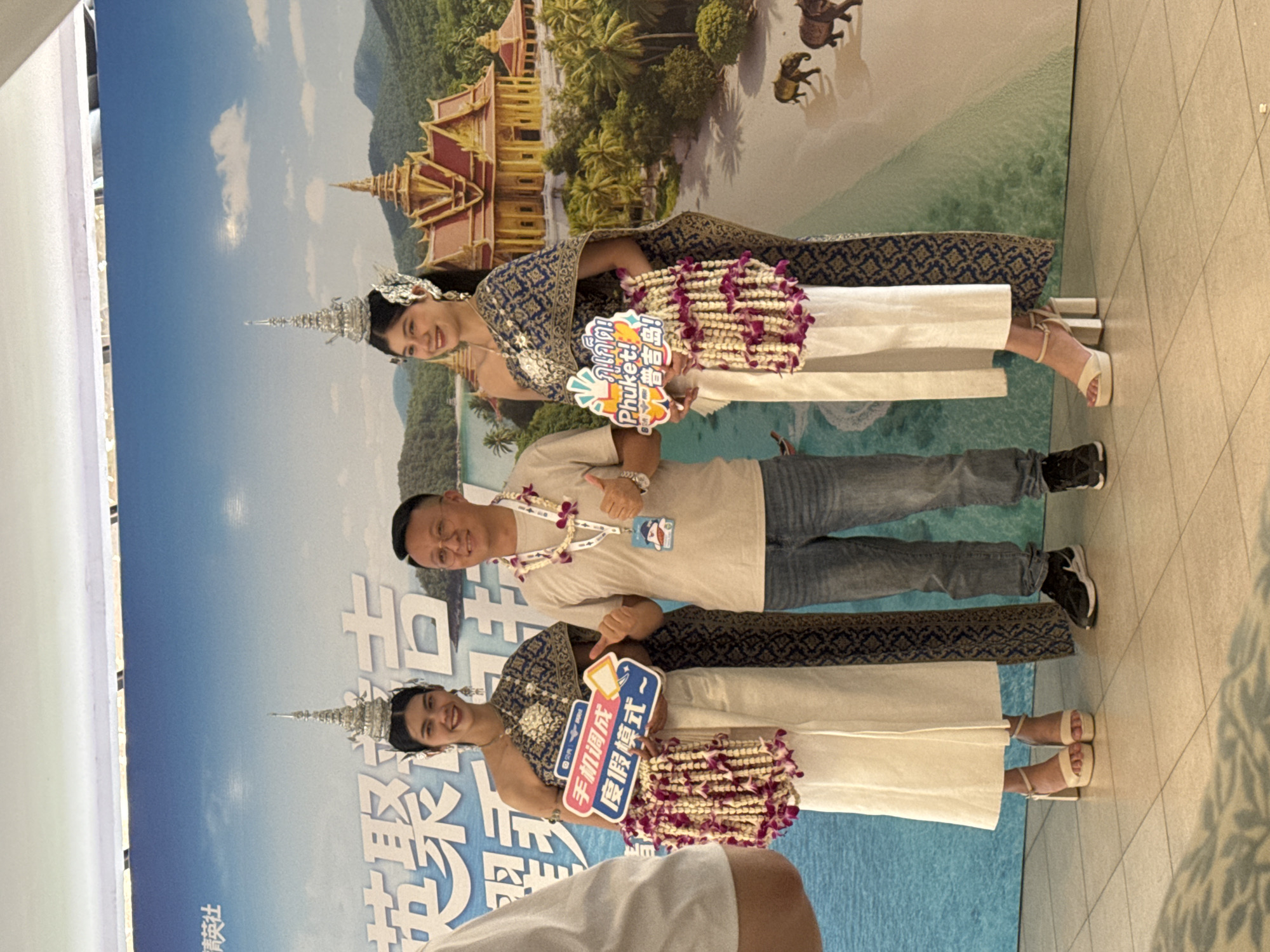 This media file captures three individuals posing in front of a large, vibrant backdrop depicting a tropical landscape. The backdrop features a clear blue sky, distant mountains, lush palm trees, a golden traditional Thai-style building, elephants, and a serene turquoise sea, strongly suggesting a destination like Phuket, Thailand.
Two women, positioned on either side, are dressed in elegant, traditional Thai-inspired outfits, including elaborate silver headpieces, patterned blue and gold sashes, and long white skirts. Both women are adorned with intricate flower garlands, some draped over their arms. A man stands between them, dressed casually in a light-colored t-shirt, blue jeans, and sneakers, also wearing a flower lei around his neck. All three subjects are smiling brightly, appearing joyful and engaged.
The women are holding promotional signboards. The sign held by the woman on the right features text in Thai ("ภูเก็ต!"), English ("Phuket!"), and Chinese ("普吉岛!"), along with the number "300". The woman on the left holds a sign displaying Chinese characters that read "手机调成 度假模式~" (Set phone to vacation mode~). Large Chinese characters, including "英聚福吉" and "精英社," are also visible on the banner behind them. The man gives a thumbs-up gesture, while the woman on the left points towards her sign. The floor in the foreground is tiled, indicating an indoor or sheltered setting, likely an airport or a tourism event, celebrating or promoting travel to Phuket, Thailand, potentially in a location like Patong.