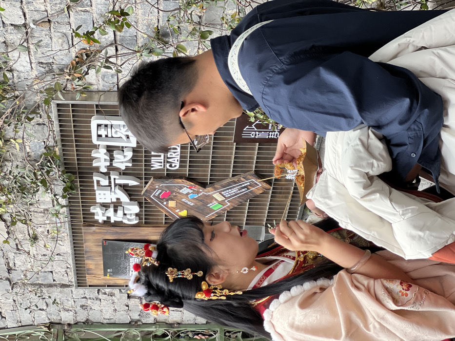 A man in a blue shirt and glasses holds out a piece of food on a stick to a woman in traditional Chinese clothing. The woman, adorned with ornate hair accessories, opens her mouth to receive the treat. They stand in front of a sign with Chinese characters, positioned near a grid and brick wall with some foliage. A map is partially visible on the sign. The man has a cream-colored jacket draped over his shoulder.