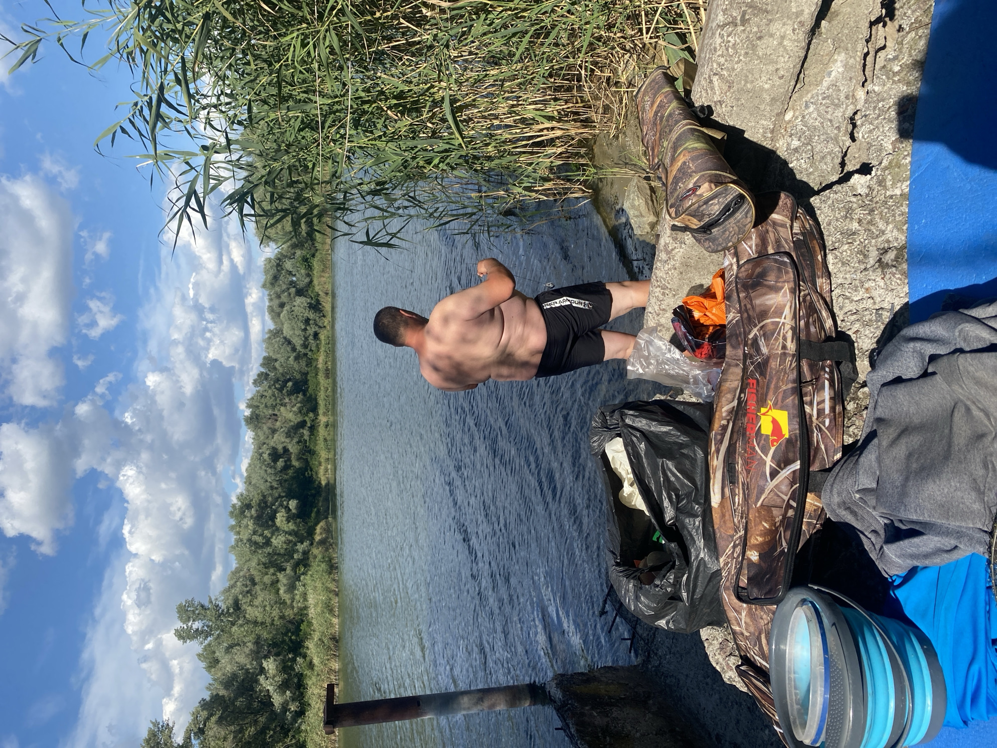 An overcast, summer day at the Dnipro River in Dnipro, Ukraine. A man, shirtless and wearing black shorts, stands in the shallow water at the river's edge, holding a fishing rod. He appears to be fishing. In the foreground, on the rocky bank, are various items associated with fishing or a day outdoors. These include a large black trash bag filled with what appears to be white material, two camouflage fishing rod bags, a foldable blue bucket, and a grey hoodie draped over a blue tarp. Dense green reeds line the riverbank, and a metal pole extends from the water to the left. The water is calm, with a light chop on the surface. In the background, a thick line of trees borders the opposite bank of the river, under a partly cloudy sky.