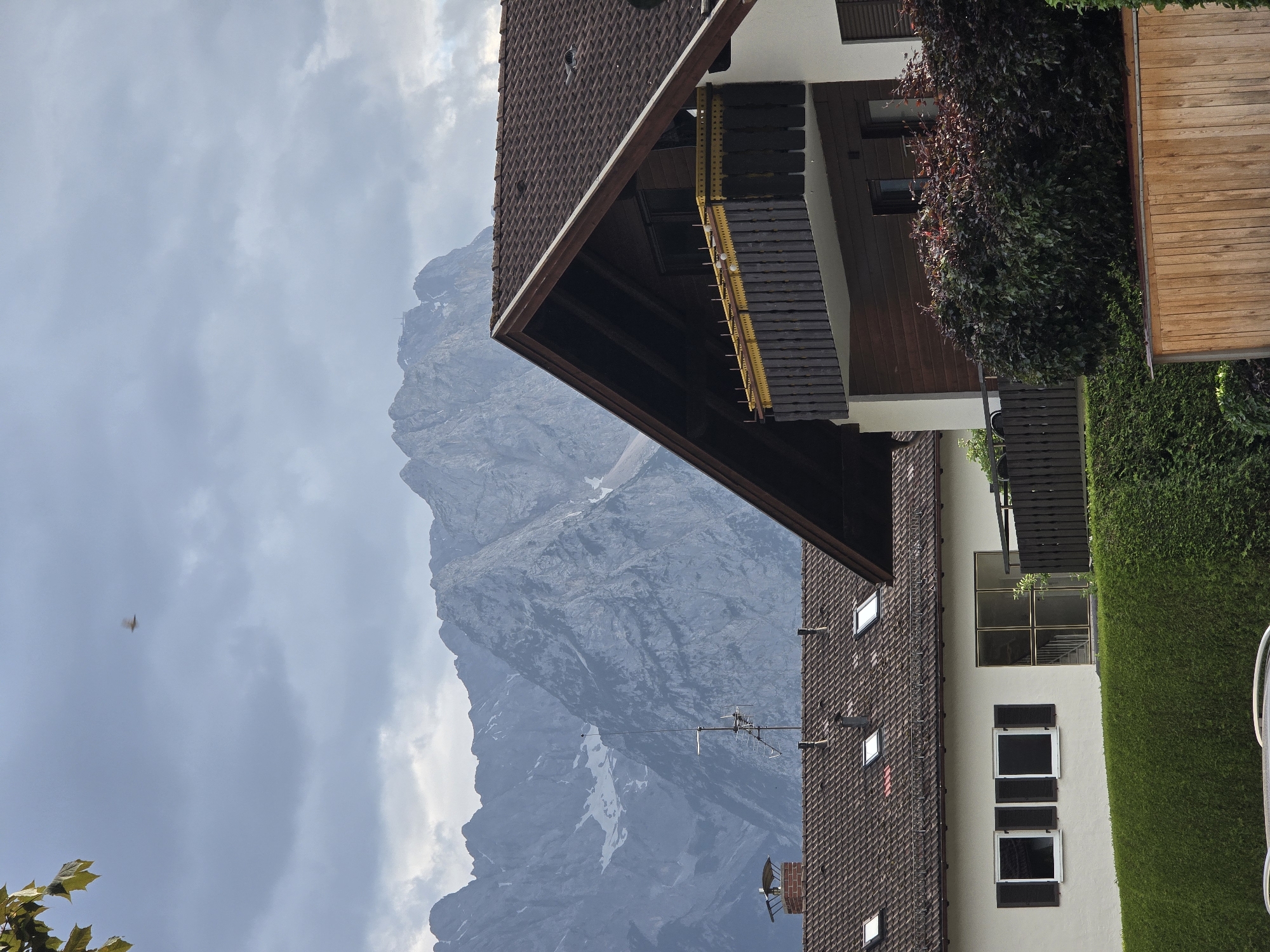 This image depicts a suburban scene in Garmisch-Partenkirchen, Germany, with a focus on residential buildings set against a dramatic mountain backdrop. The foreground is dominated by the rooftops and upper stories of two houses. The house on the right features a prominent dark brown tiled roof with a steeply pitched gable. It has a wooden-paneled upper story and a balcony with dark vertical slats and a yellow decorative trim. Below the balcony, dark red-leaved bushes create a dense hedge. The house on the left has a similar tiled roof and a lighter colored exterior wall. It features several rectangular windows with dark shutters and a large glass door leading to what appears to be an interior staircase. Both houses are separated by neatly trimmed green hedges.
In the middle ground, the roofs of the houses are visible, with a chimney on the left house and an antenna array on the roof of the house on the left. Beyond the houses, a towering mountain range with rugged, rocky peaks dominates the background. Some of the higher elevations of the mountains are dusted with snow. The sky is overcast with heavy, gray clouds, creating a somewhat somber atmosphere. A single bird is visible in flight against the cloudy sky. The overall impression is one of a dwelling in a scenic, mountainous region.