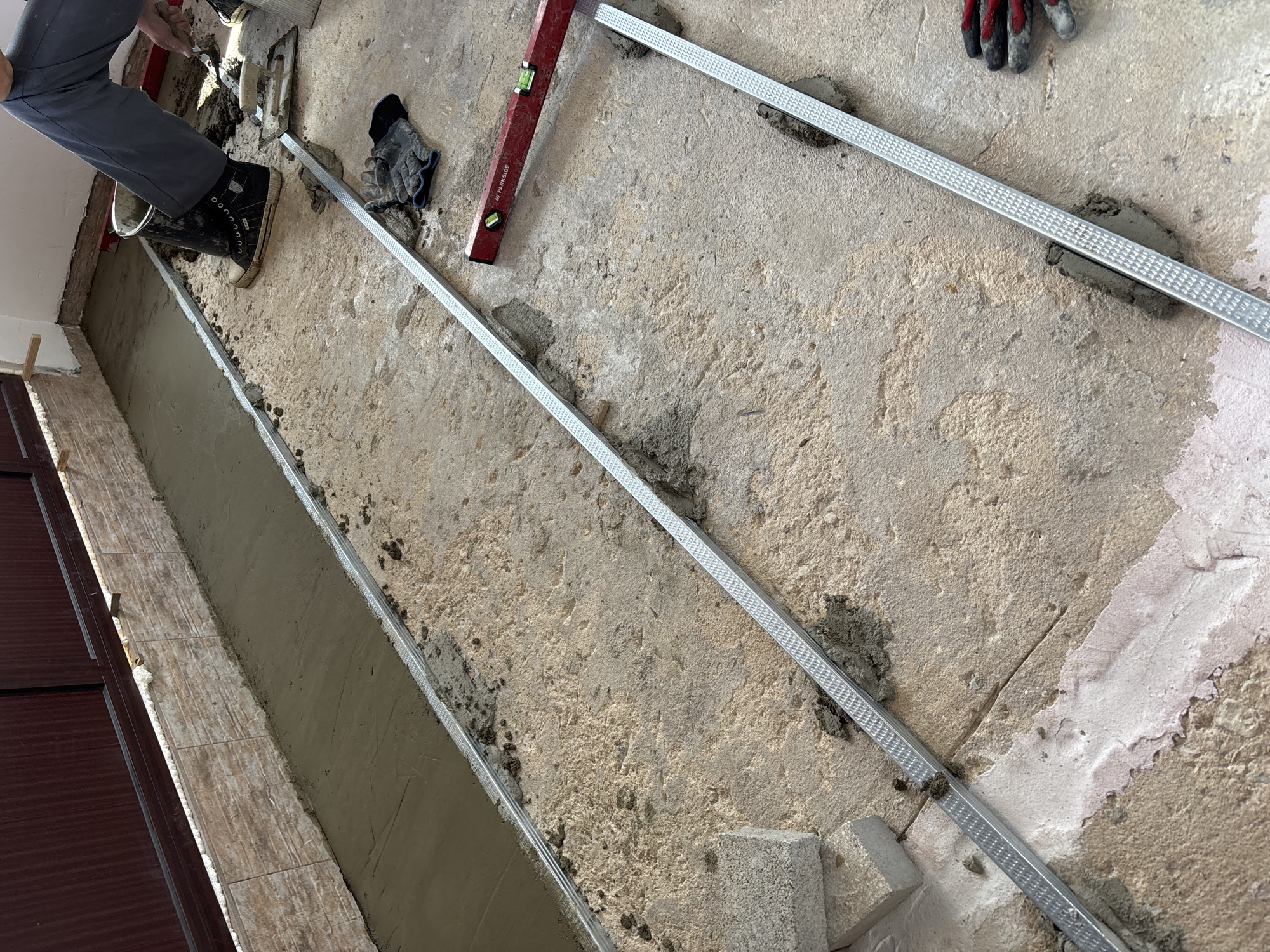 The scene shows a construction or renovation project underway, likely indoors on a concrete floor in Pchelnik, Bulgaria. A worker is visible, wearing gray trousers and black work boots, with a bucket nearby. Various tools and materials are scattered around, including metal profiles, a level, a trowel, gloves, and bricks. Fresh mortar or cement is being applied along the edge of a tiled section. The metal profiles are being used as guides for the application of the mortar.