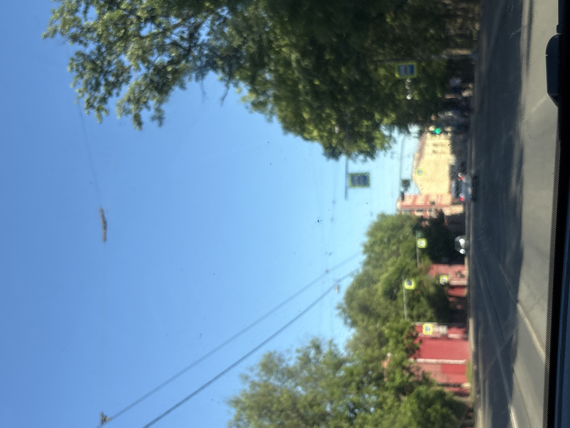 The image captures a wide urban thoroughfare in Saint Petersburg, Russia, observed from inside a vehicle through a somewhat dusty windshield under a clear, bright blue sky. A sparse scattering of small dark specks, likely debris or insects, is visible on the windshield surface.
Numerous overhead wires, part of an electrical network for public transport such as trams or trolleybuses, stretch across the sky. These wires are supported by poles and insulators, and one section shows a complex junction of wires.
The road surface is asphalt, featuring distinct tram tracks running down its center. The street appears to be multi-laned, receding into a blurry distance where the light casts long shadows.
On the left side of the street, a continuous row of multi-story buildings, primarily painted in deep red hues, is visible behind a dense line of green trees. These buildings exhibit a utilitarian architectural style, common in late 19th and early 20th-century Russian urban development. Several traffic signals with characteristic yellow frames, including a blue pedestrian crossing sign with a white figure, are mounted above the trees. A dark-colored vehicle is partially obscured by the trees on this side.
On the right side of the street, another line of lush green trees partially obscures the buildings behind them. Further along, a lighter-colored, possibly yellow or ochre, building with classical architectural details is visible. A green traffic light is active, and several blurry human figures, presumably pedestrians, are discernible on the sidewalk near it. A light-colored car is seen driving away in the distance on this side.
The scene depicts a typical major avenue in Saint Petersburg, characterized by its combination of historic and early 20th-century architecture, prominent green spaces, and integrated electric public transport infrastructure.