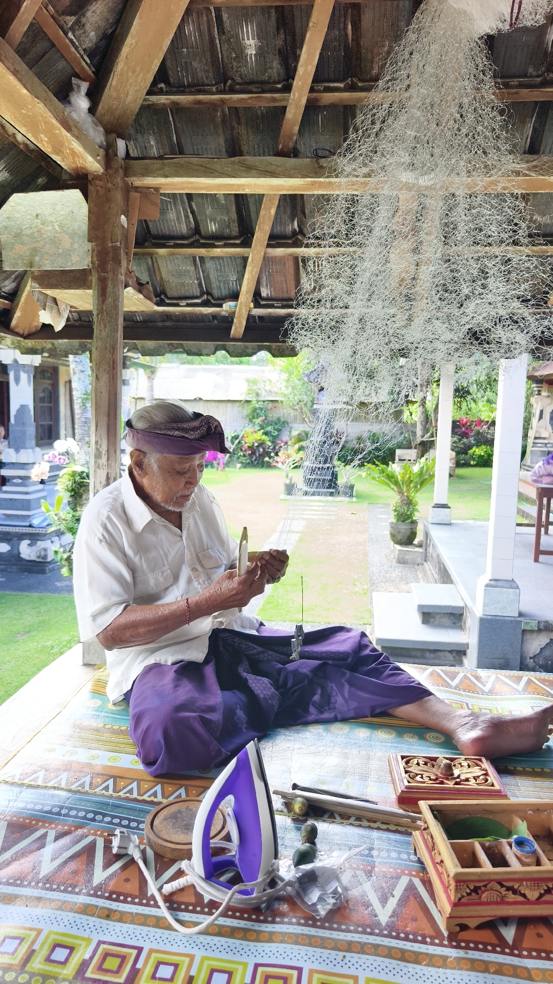An elderly Balinese man is seated in a covered outdoor space, possibly a pavilion or veranda, in Pecatu, Indonesia. He wears a white short-sleeved shirt, purple pants, and a traditional Balinese headdress. He is focused on a task, holding a small wooden instrument in his hands, seemingly weaving or crafting something with fine white threads that are suspended from the ceiling above.
The seating area features a colorful patterned surface, and various objects are arranged around him, including a purple and white iron, wooden boxes, small wooden sticks, and a few green objects. In the background, there is a view of a garden with lush greenery, flowers, and a small fountain or water feature. Part of a traditional Balinese building is also visible in the background. The space above him is covered by a wooden roof structure with an intricate network of beams and rafters, from which the white threads are suspended.