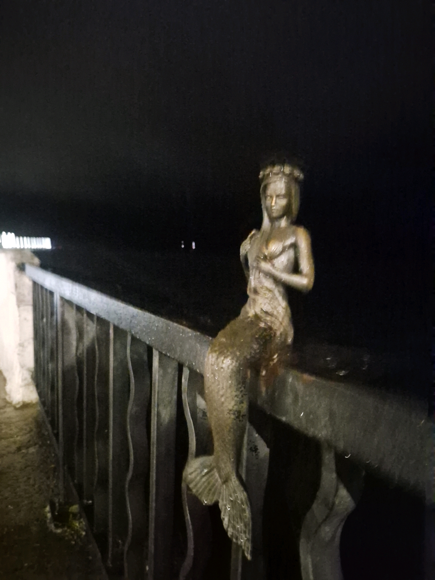This is a nighttime, medium-shot photograph of a bronze statue of a mermaid sitting on the railing of a bridge. The statue is detailed, with the mermaid having long hair, a crown, and a tail. The bridge railing is made of dark metal with vertical wavy bars. To the left of the railing, there is a stone embankment. The background is dark, indicating nighttime, with some distant lights visible in the water. The statue is likely located in Chaikovsky, Russia, near the Kama River, as indicated by the general context.