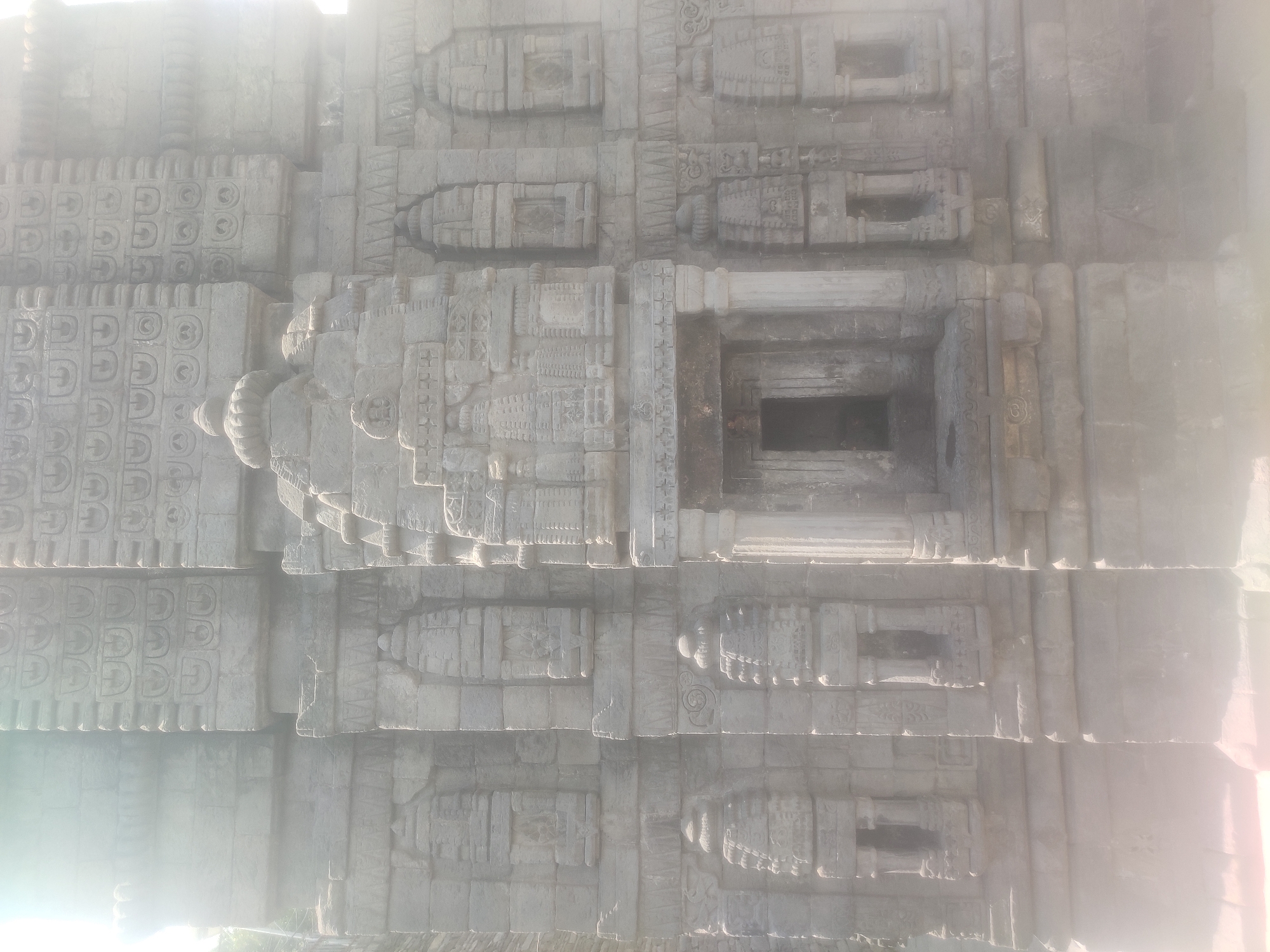 The image shows the exterior of an ancient stone temple, likely located in Naggar, India. The temple's facade is intricately carved with architectural details, including decorative niches, pillars, and what appear to be miniature temple structures. The stonework is weathered, suggesting age. Sunlight is casting a bright glare, particularly on the left side of the image, which obscures some of the finer details. The overall impression is one of historical and religious significance.