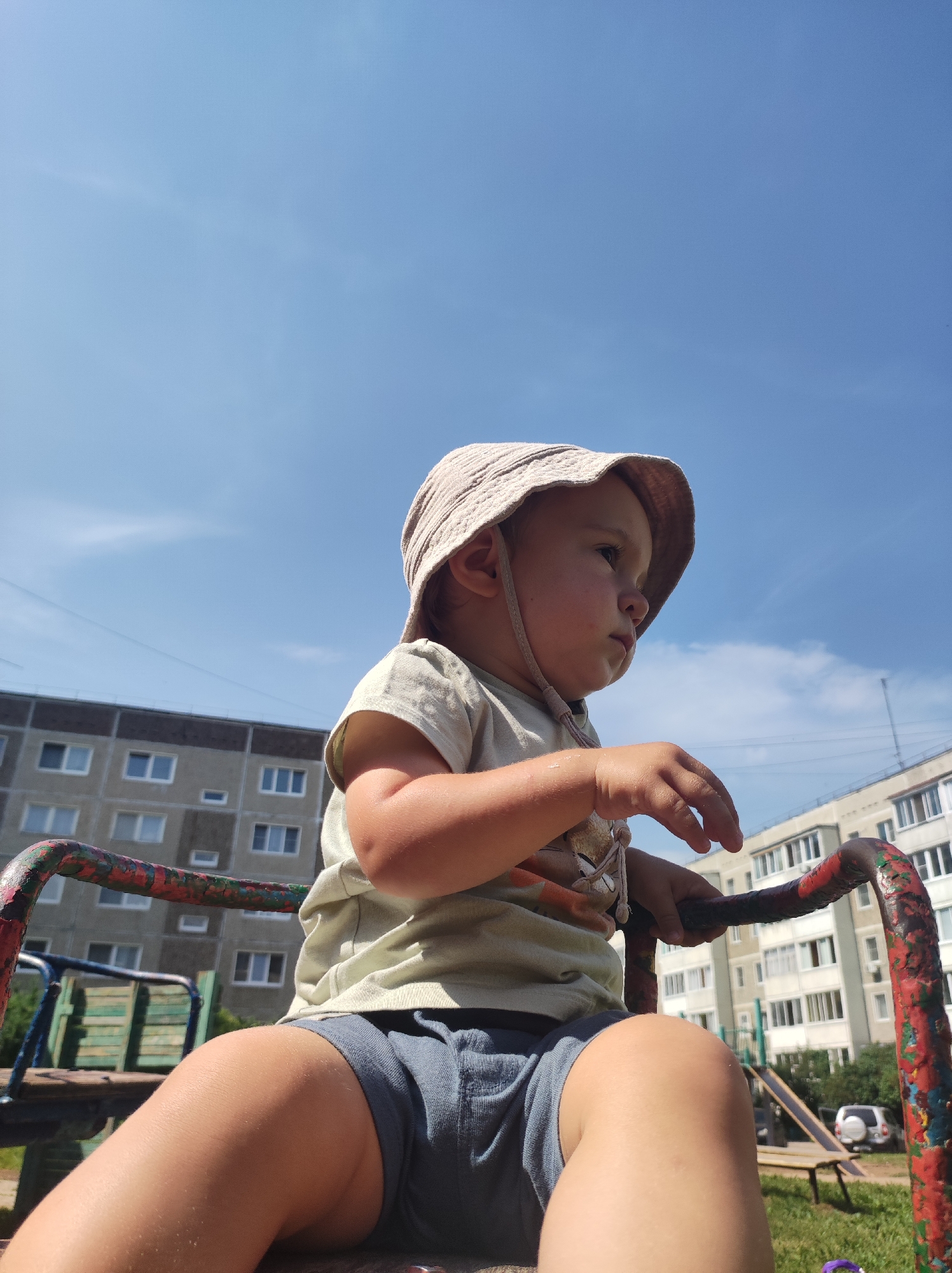 An eye-level, medium shot captures a young child, wearing a light-colored bucket hat and a pale green t-shirt with a graphic print, sitting on a rusty red and green metal bar, likely part of a playground ride. The child is facing to the right, with their gaze directed off-camera. Their small hands are resting on the metal bar. The child's legs are bent at the knee, wearing dark gray shorts. In the background, a tall, grey apartment building with numerous windows is visible, along with another lighter-colored apartment building further away. The sky is a clear blue with a few wispy clouds. The scene appears to be an outdoor playground in an urban setting. The image is taken from a low angle, looking up at the child. The overall impression is of a sunny day.