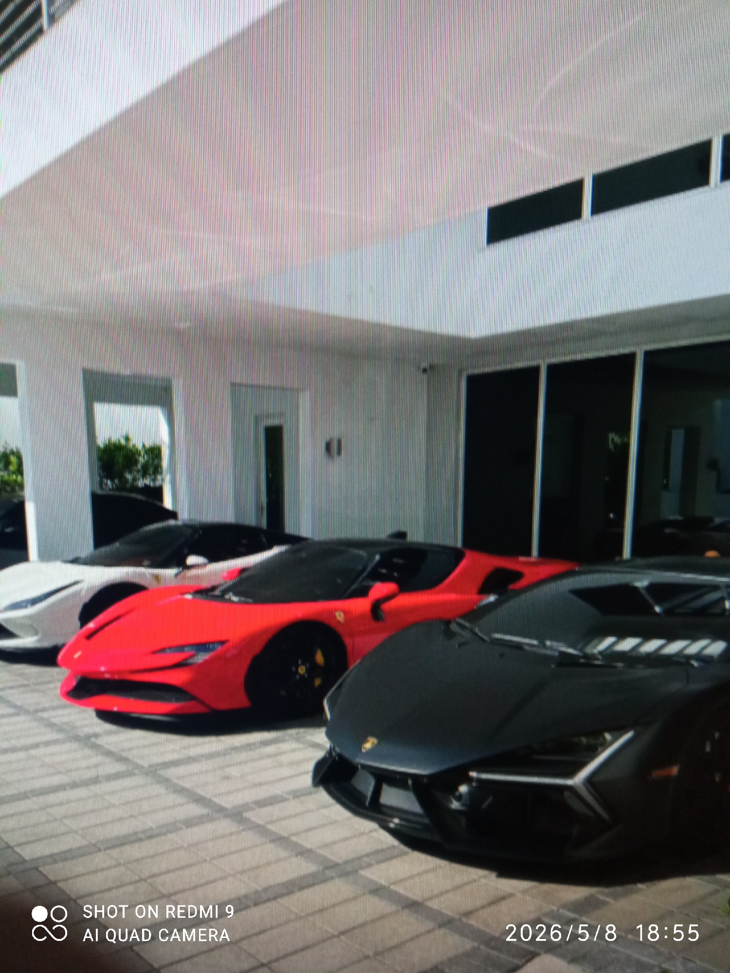 The image depicts a row of luxury sports cars parked outside a modern building in an unidentified location. From left to right, there is a white car, a red Ferrari SF90 Stradale, and a black Lamborghini Aventador. The cars are parked on a light gray paved surface. The building behind the cars is white with a modern design, featuring rectangular windows and columns. The photo was taken on May 8, 2026 at 18:55 with a Redmi 9 AI Quad camera.