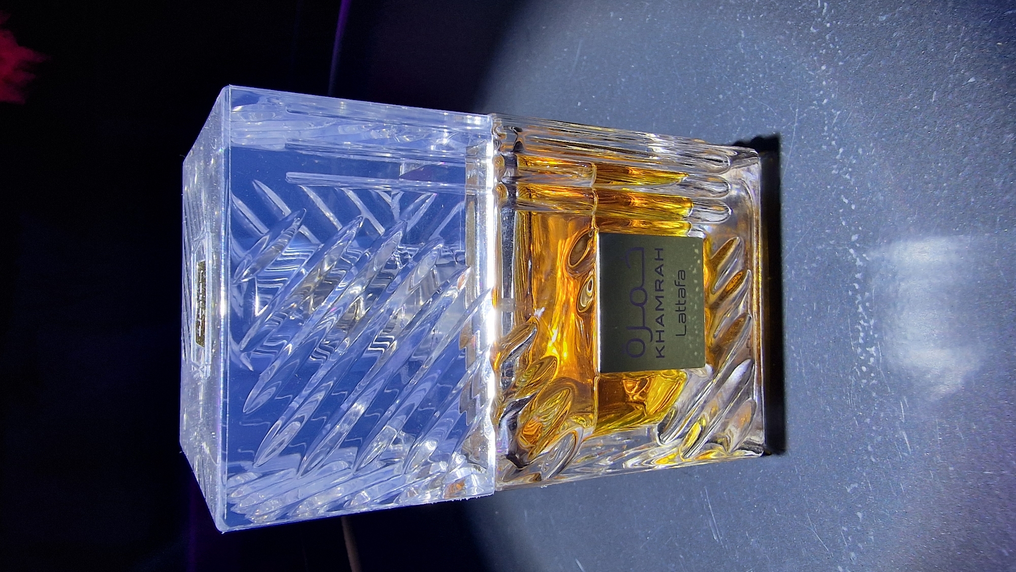 The image shows a bottle of Khamrah Lattafa perfume. The bottle is rectangular with a clear, elaborately cut glass cap on the left and amber-colored perfume within the right side. A green label on the right side displays the perfume's name in Arabic and English. The bottle sits on a reflective, dark gray surface, with a dark background partially visible. The image was not taken in Baan Bang Rak, Thailand.