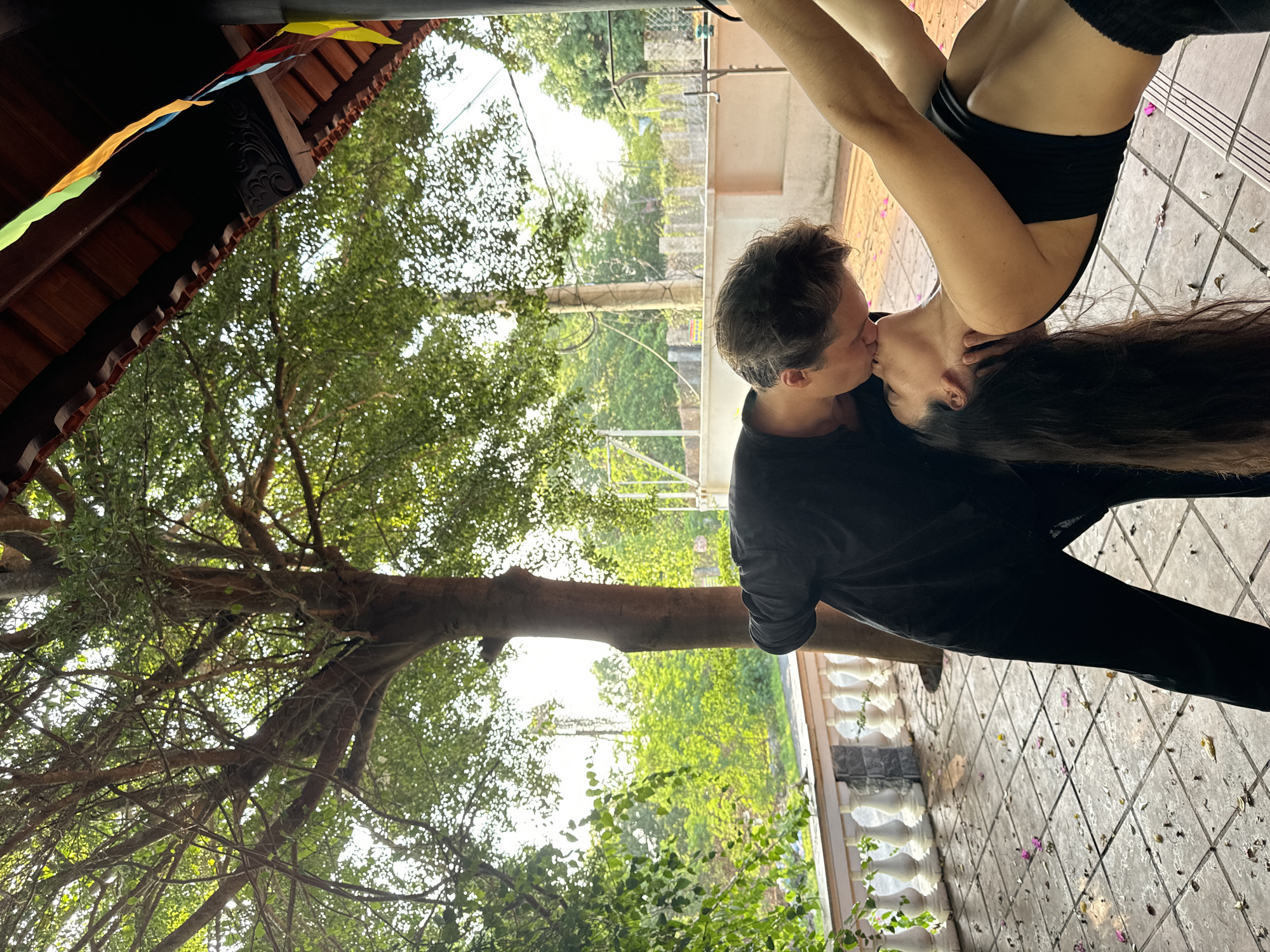 This image captures an intimate interaction between two individuals on an outdoor tiled patio in Mũi Né, Vietnam. A male, dressed in a black long-sleeved shirt and black pants, is positioned leaning forward, his hands near the female's head. A female, with long dark hair, wears a black sports bra and dark bottoms, performing a backward arching pose while being kissed by the male. The patio floor is covered with square, light-colored tiles, appearing wet with scattered pink petals and green leaves. A white balustrade with ornate balusters borders the patio on the left. The background features a large tree with a prominent trunk and dense green canopy. Beyond this, lush green vegetation and a distant tall, slender structure are visible under a bright sky. The upper right corner reveals a traditional roof structure with red tiles, carved wooden supports, and colorful fabric bunting hanging from it.